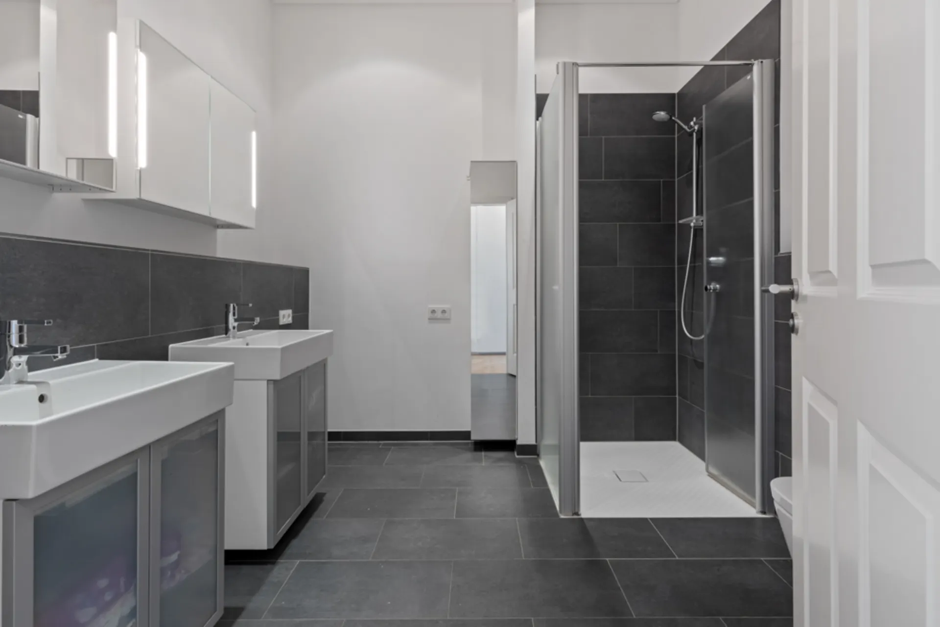 Modernes Badezimmer mit zwei Waschbecken und einer Dusche mit Glaswand und dunklen Fliesen.