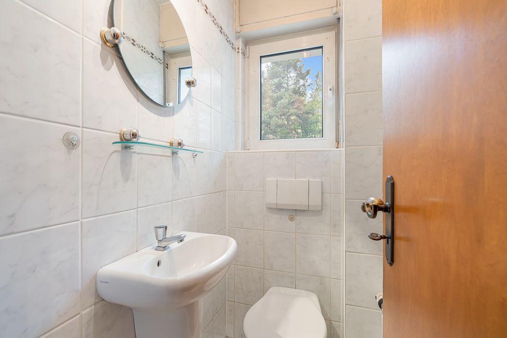 Kleines Bad mit Waschbecken, Toilette, Spiegel und Fenster, weiße Fliesen an den Wänden.