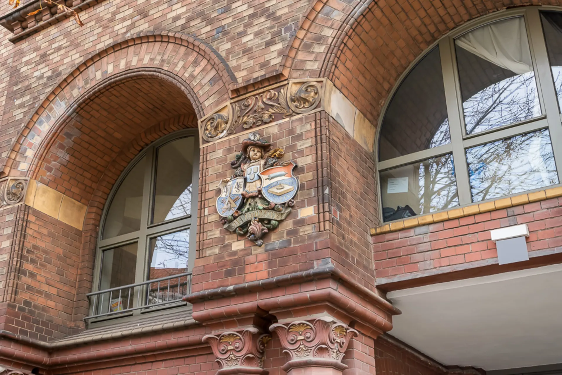 Backsteinfassade mit zwei großen Rundbogenfenstern und einem bunten Wappenrelief zwischen den Fenstern.