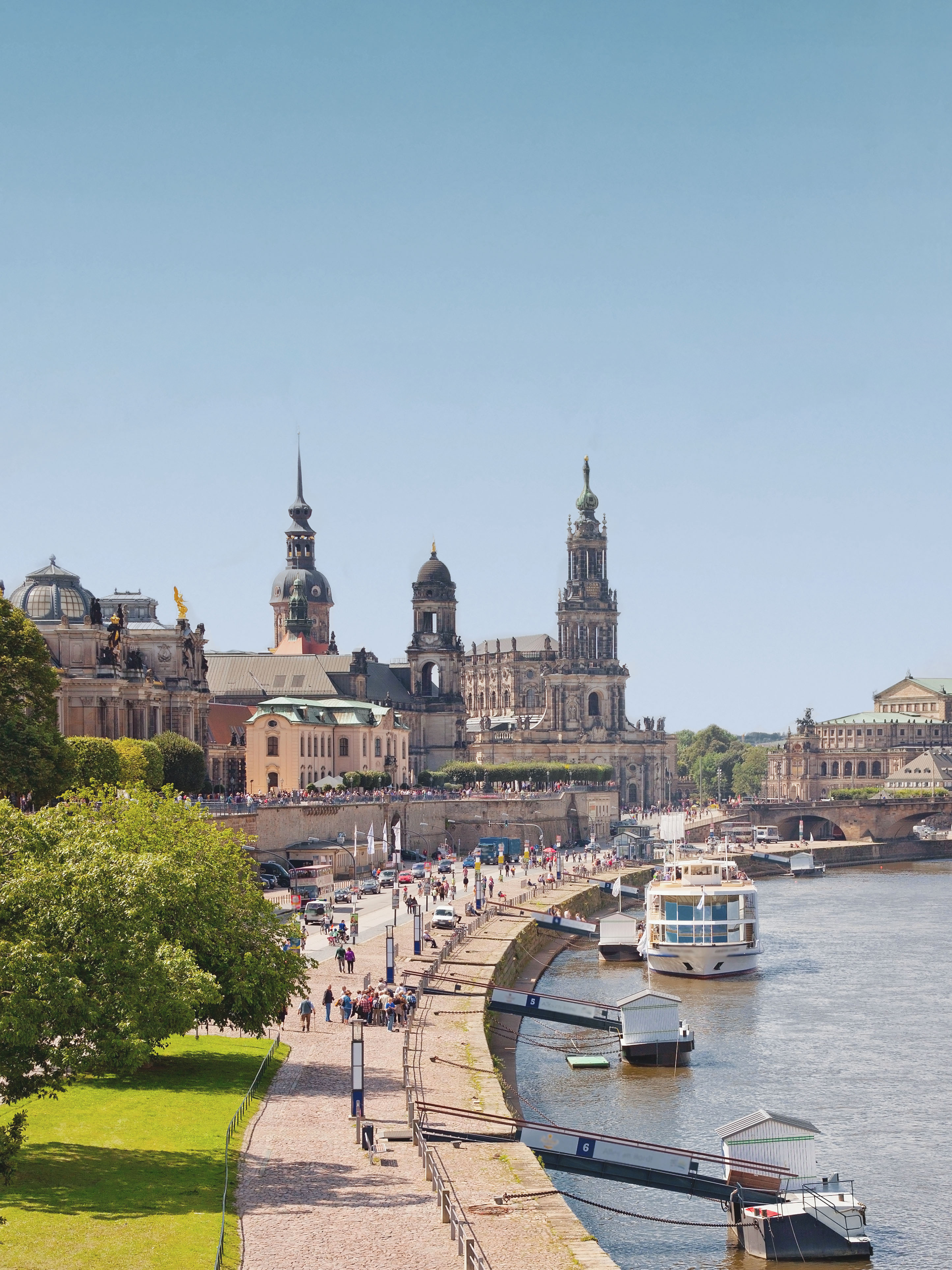 Promenade mit Menschen entlang eines Flusses, Boote und historische Gebäude im Hintergrund.