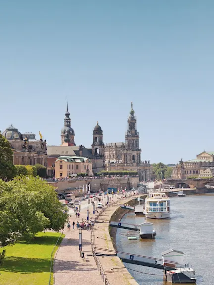Promenade mit Menschen entlang eines Flusses, Boote und historische Gebäude im Hintergrund.