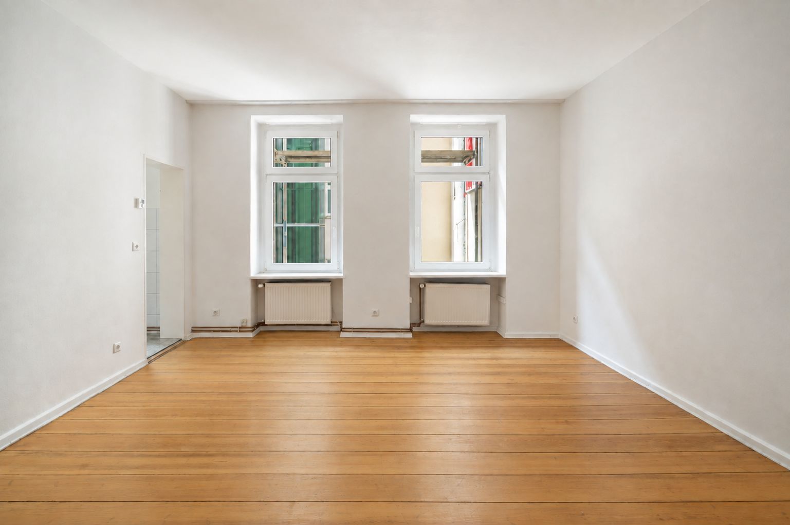 Helles, leeres Zimmer mit Holzboden und zwei Fenstern an der Rückwand.