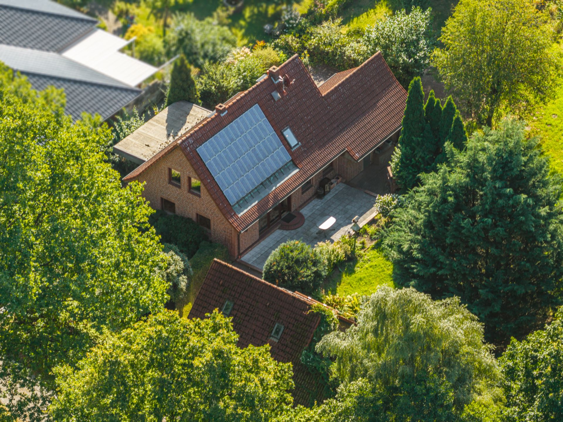 Luftaufnahme eines Einfamilienhauses mit Solarpanel auf dem Dach und Terrasse, umgeben von Bäumen.