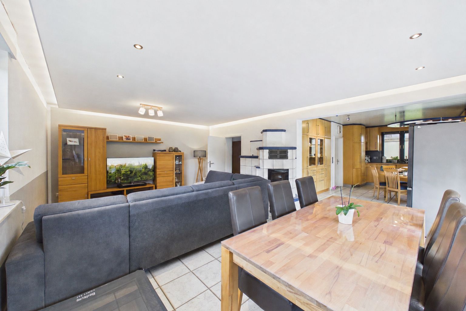 Wohnzimmer mit grauem Sofa, Esstisch, Holzschränken und offener Küche im Hintergrund.