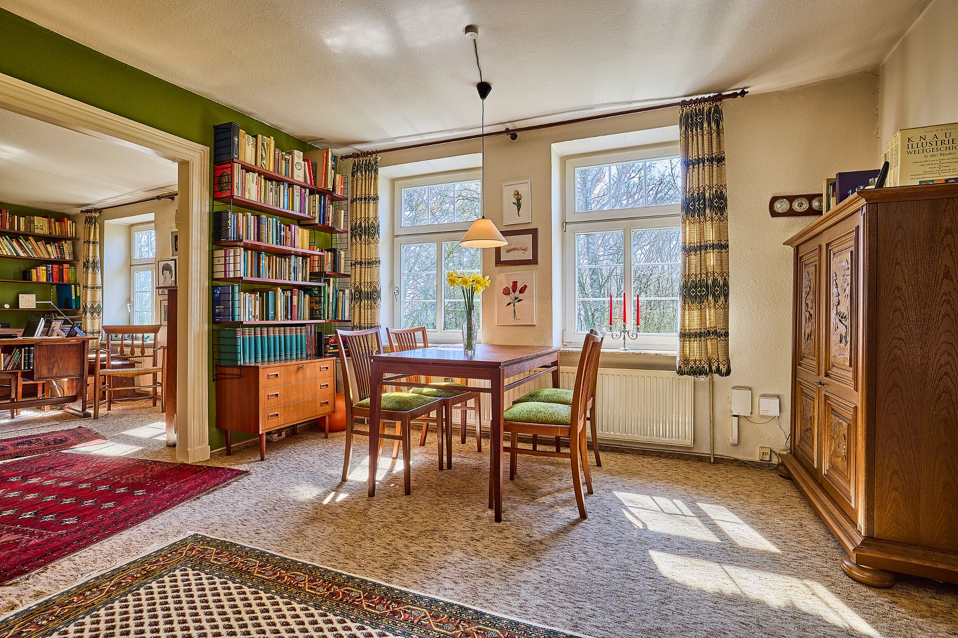 Gemütlicher Wohnraum mit Esstisch, vier Stühlen, Bücherregalen und großen Fenstern.