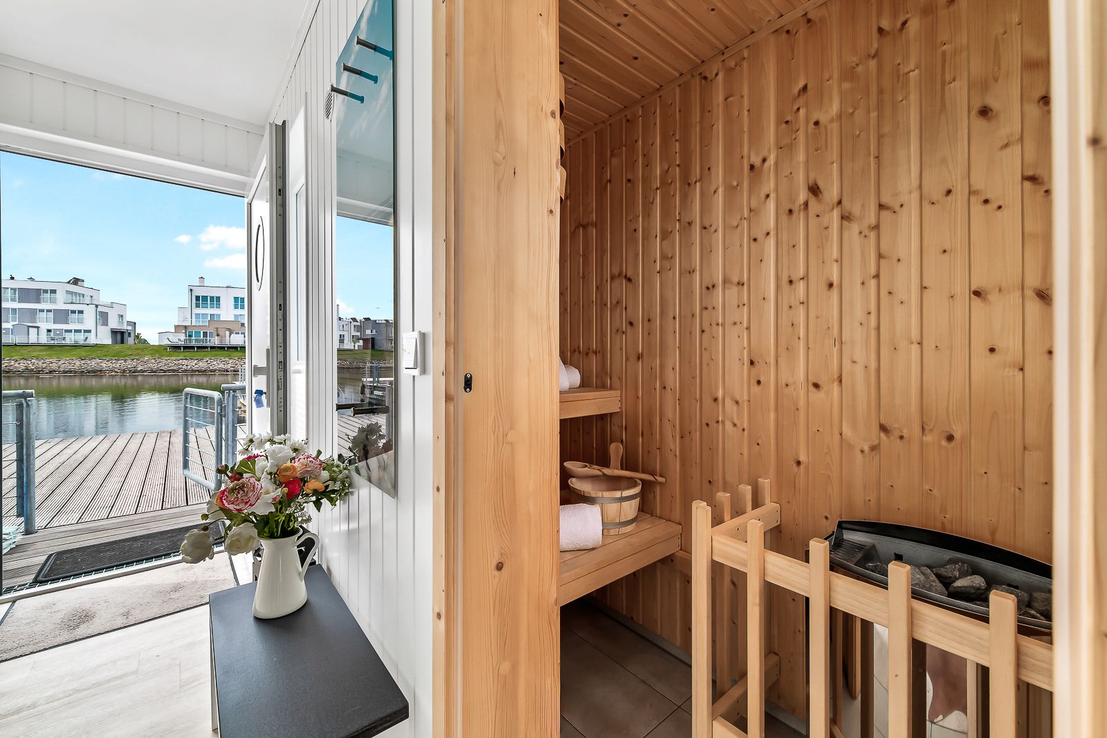 Holzsauna mit zwei Bänken neben einem offenen Eingang mit Blick auf Wasser und moderne Häuser.