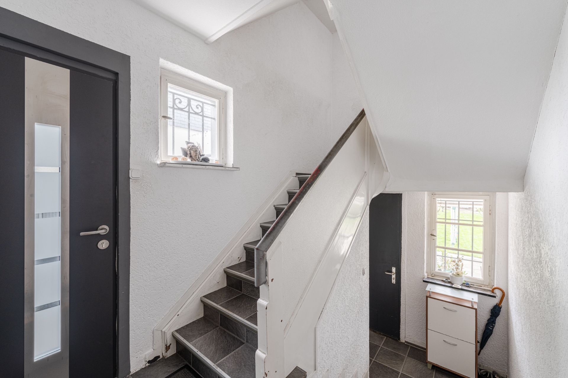 Heller Flur mit Treppe, zwei Fenstern, schwarzer Tür und kleinem Schrank mit Regenschirm.