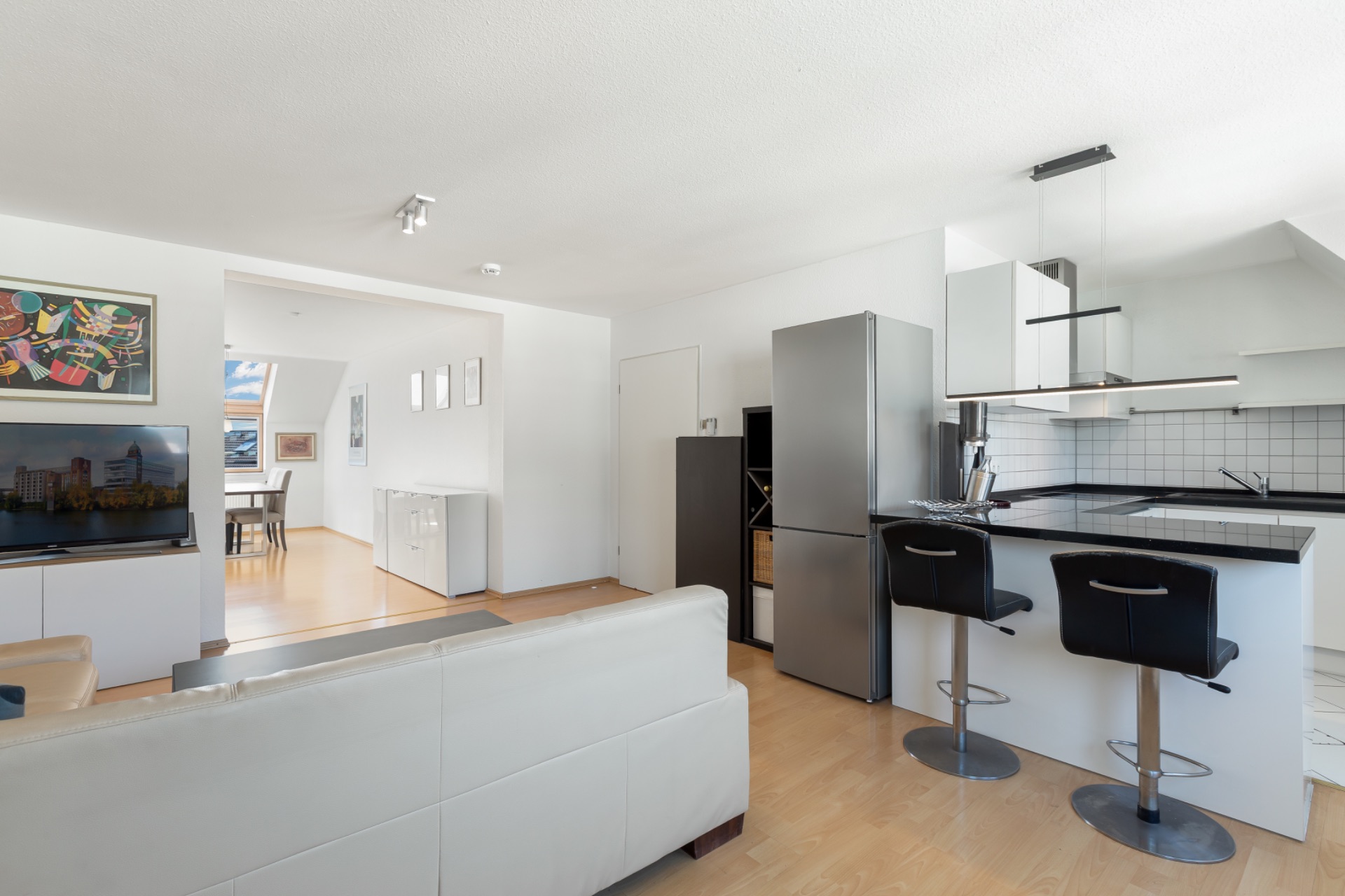 Helles Wohnzimmer mit offener Küche, Sofa, zwei Barhockern und Esstisch im Hintergrund.
