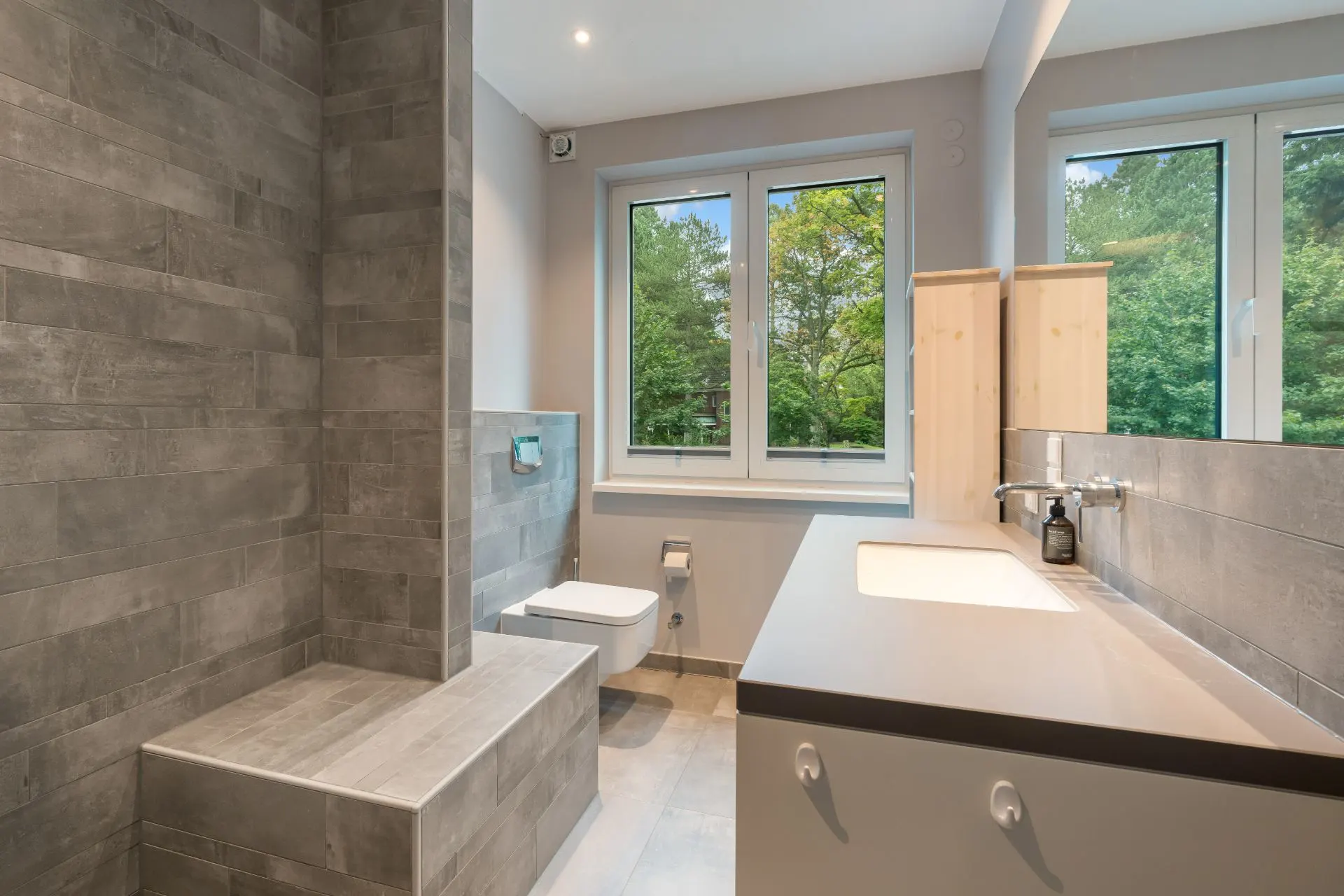 Modernes Badezimmer mit grauen Fliesen, großem Fenster, Waschbecken und wandhängender Toilette.