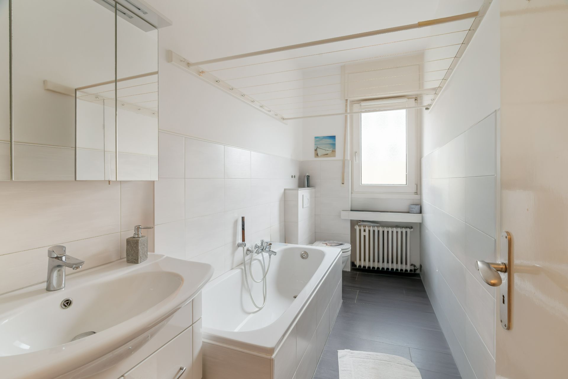 Helles Badezimmer mit Badewanne, Waschbecken, Spiegel, Fenster und Heizung.
