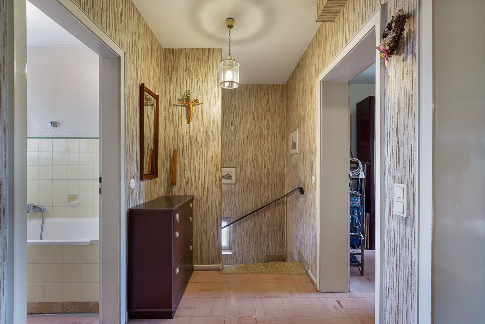 Flur mit Treppe nach unten, Kommode, Spiegel, Zugang zu Bad und weiterem Zimmer.
