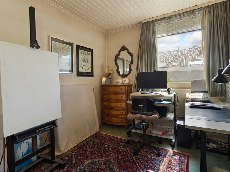 Arbeitszimmer mit Schreibtisch, Computer, Staffelei und Kommode, großes Fenster mit Vorhängen.