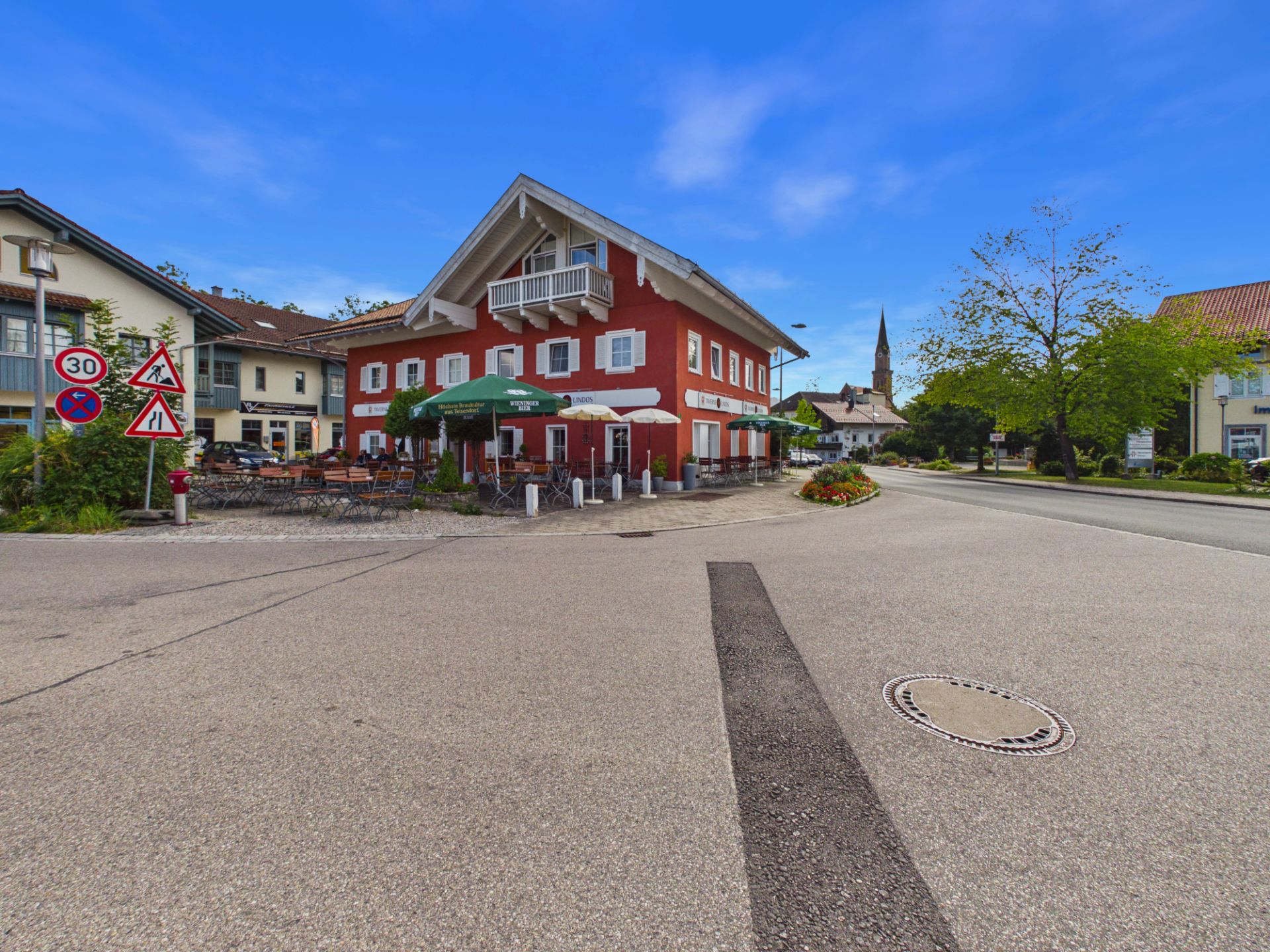 Rotes Wohngebäude mit Balkon und Terrasse an einer Straßenecke, draußen Sitzgelegenheiten und Schilder.