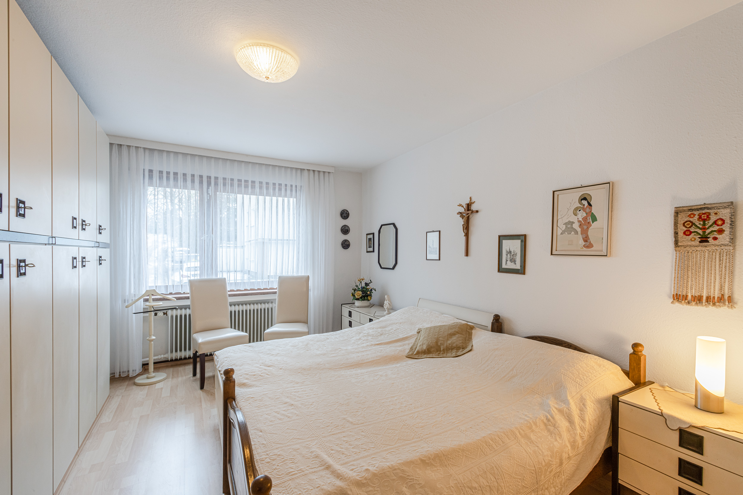 Helles Schlafzimmer mit Doppelbett, großem Schrank, zwei Stühlen am Fenster und Bildern an der Wand.