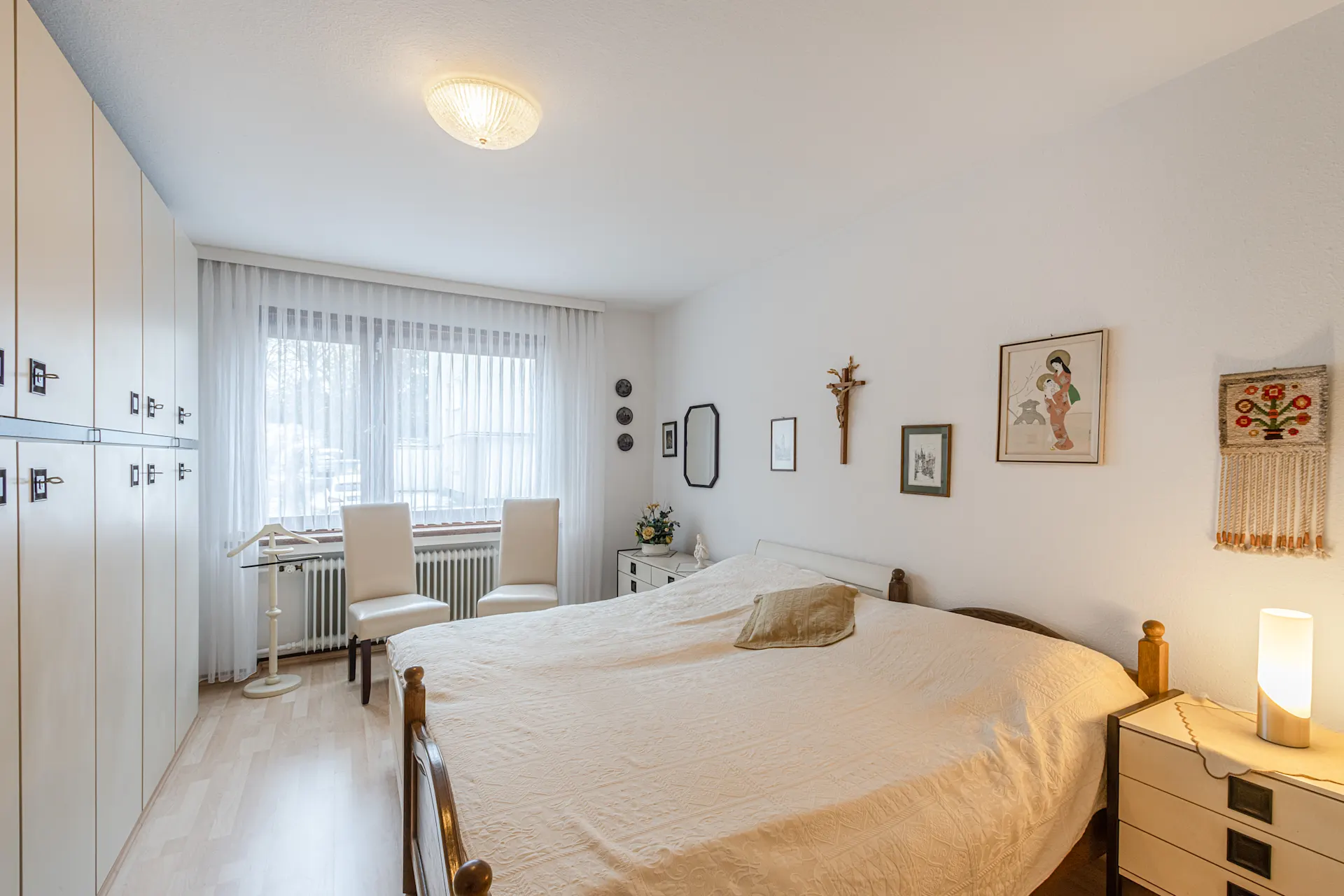 Helles Schlafzimmer mit Doppelbett, großem Schrank, zwei Stühlen am Fenster und Bildern an der Wand.