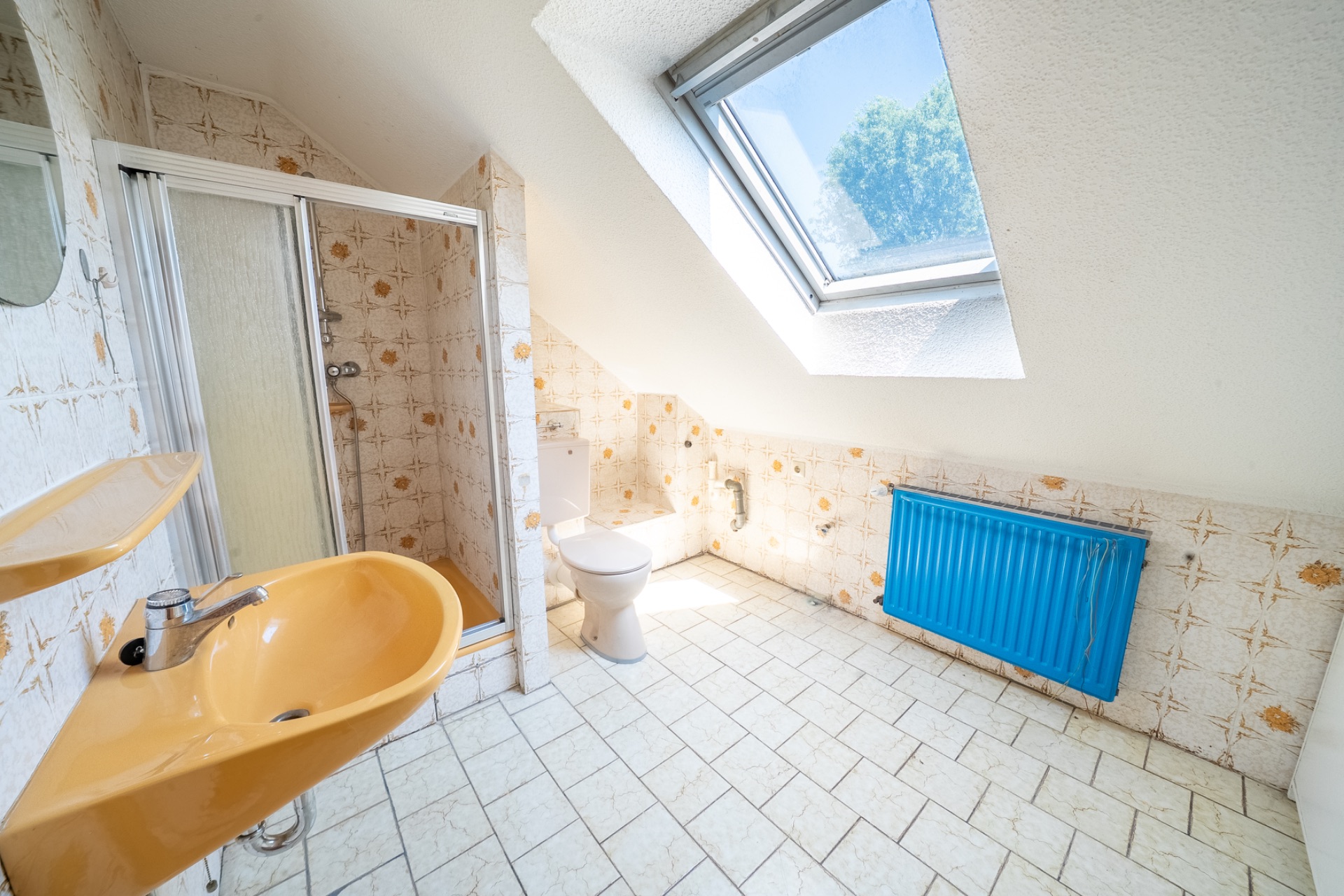 Helles Badezimmer mit gelbem Waschbecken, Dusche, WC, blauem Heizkörper und Dachfenster.