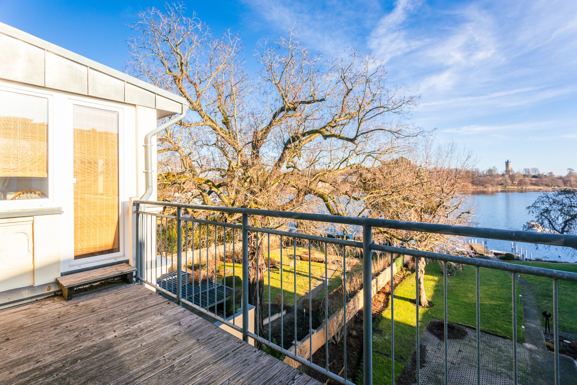 Balkon mit Holzfußboden, Blick auf Garten, Bäume und einen See im Hintergrund.