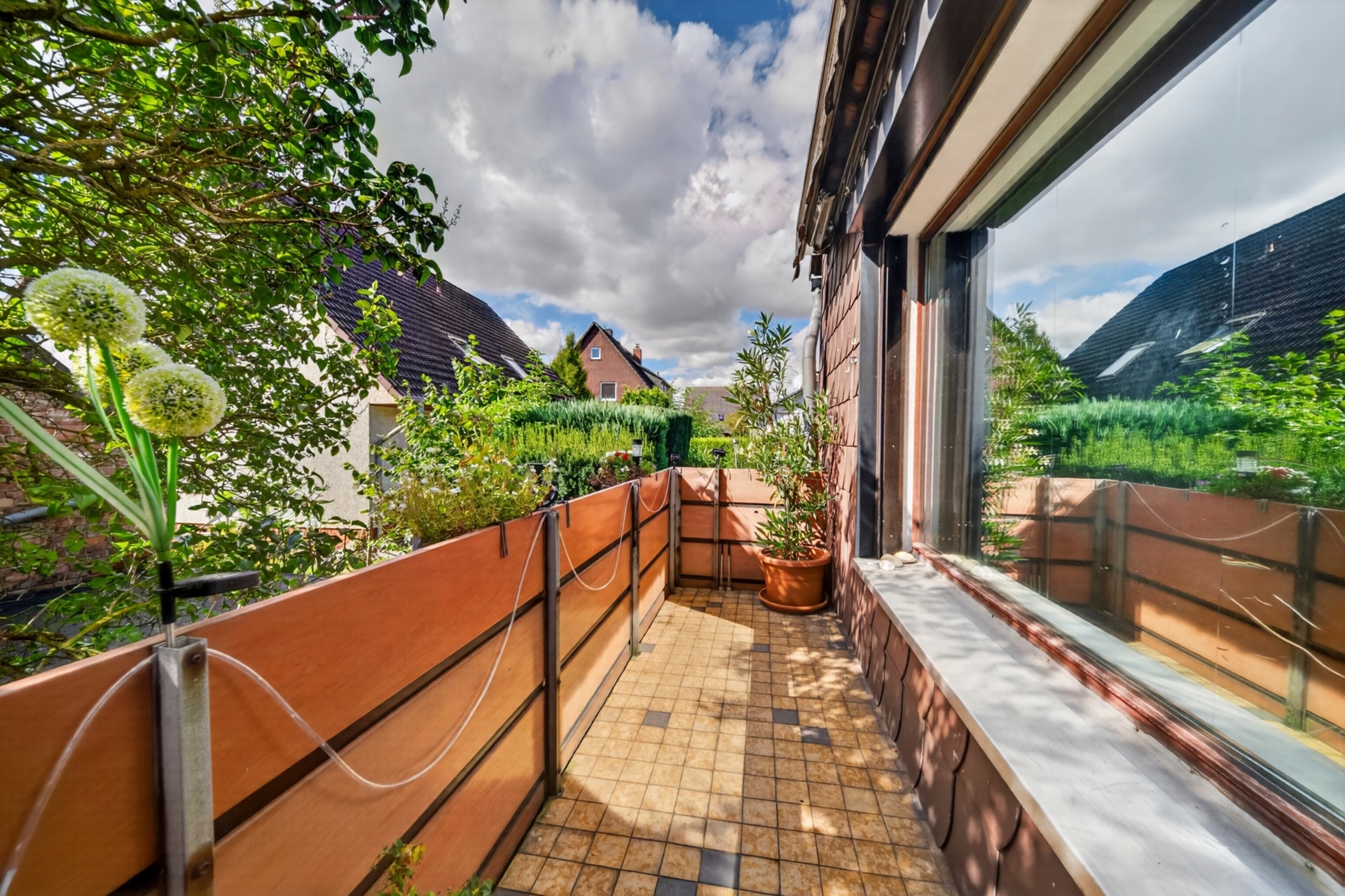 Balkon mit Pflanzen, Fliesenboden und Blick auf Nachbarhäuser an einem sonnigen Tag.