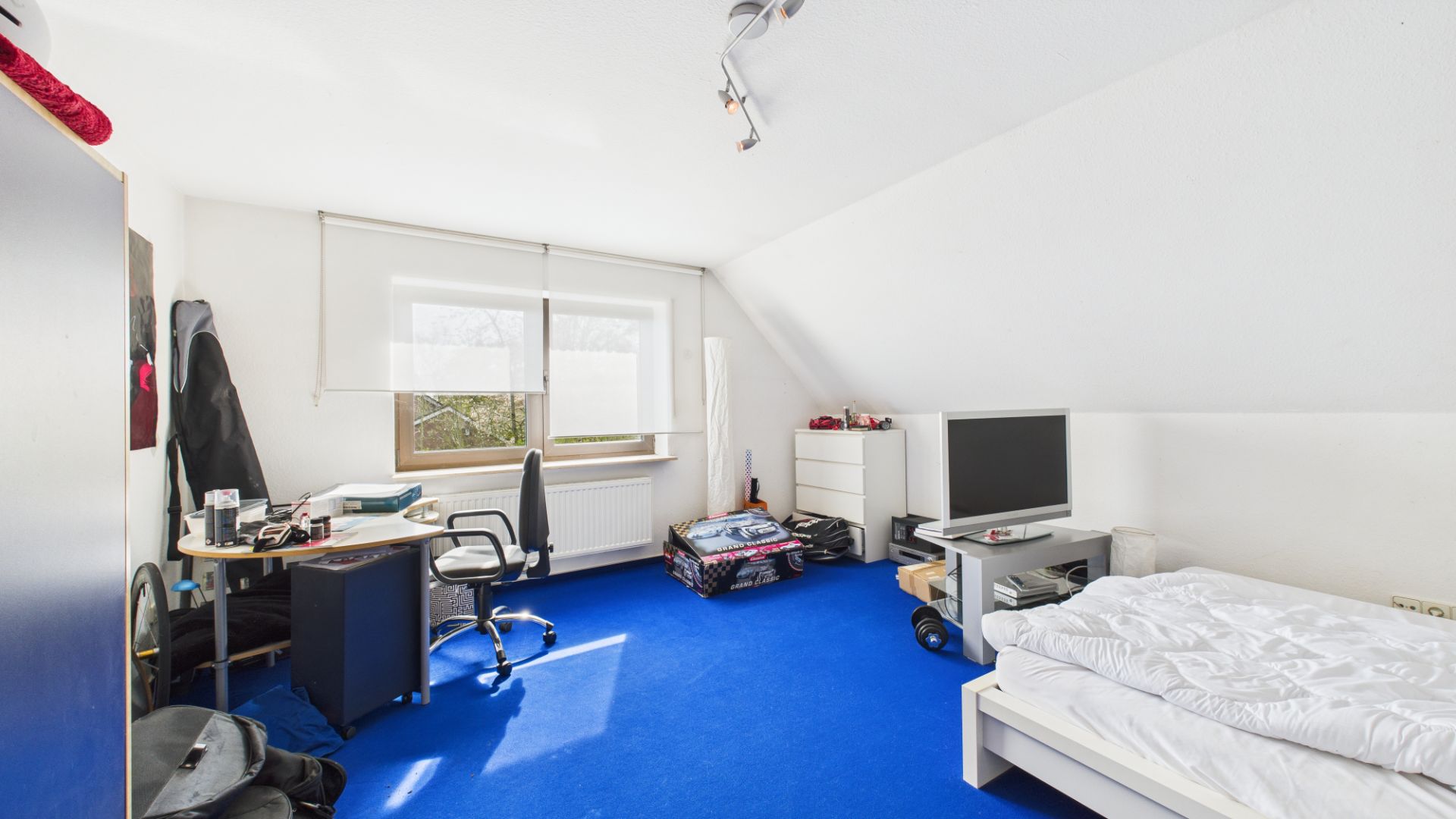 Helles Schlafzimmer mit blauem Teppich, Bett, Schreibtisch und Fernseher unter Dachschräge.