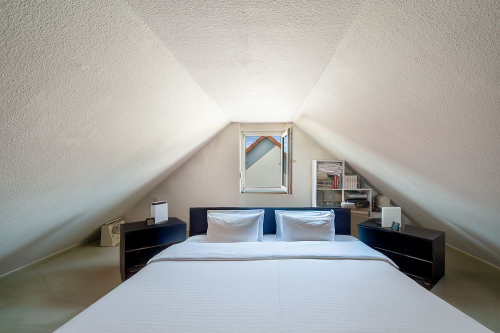 Dachzimmer mit Doppelbett, zwei Nachttischen und einem kleinen Fenster in der Mitte.