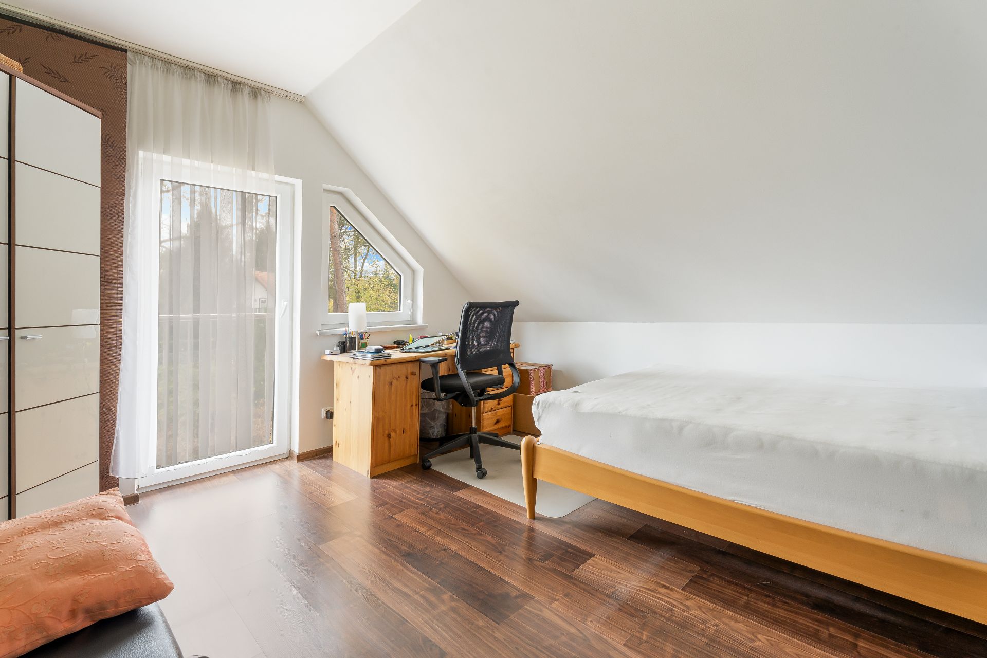 Helles Zimmer mit Holzboden, Bett, Schreibtisch, Bürostuhl und Fenstern unter Dachschräge.