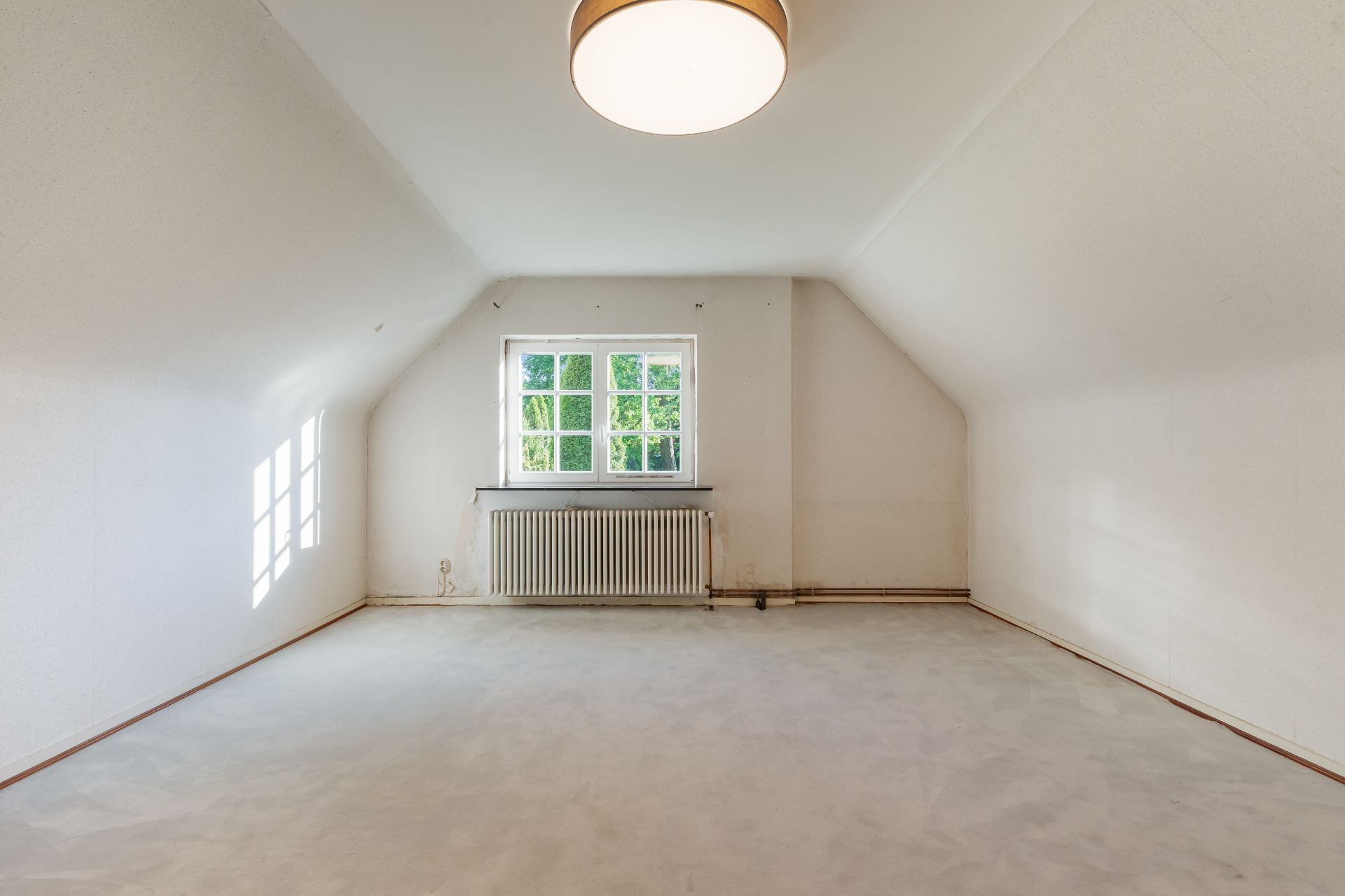 Helles, leeres Zimmer mit Dachschrägen, Fenster, Heizkörper und Deckenlampe.