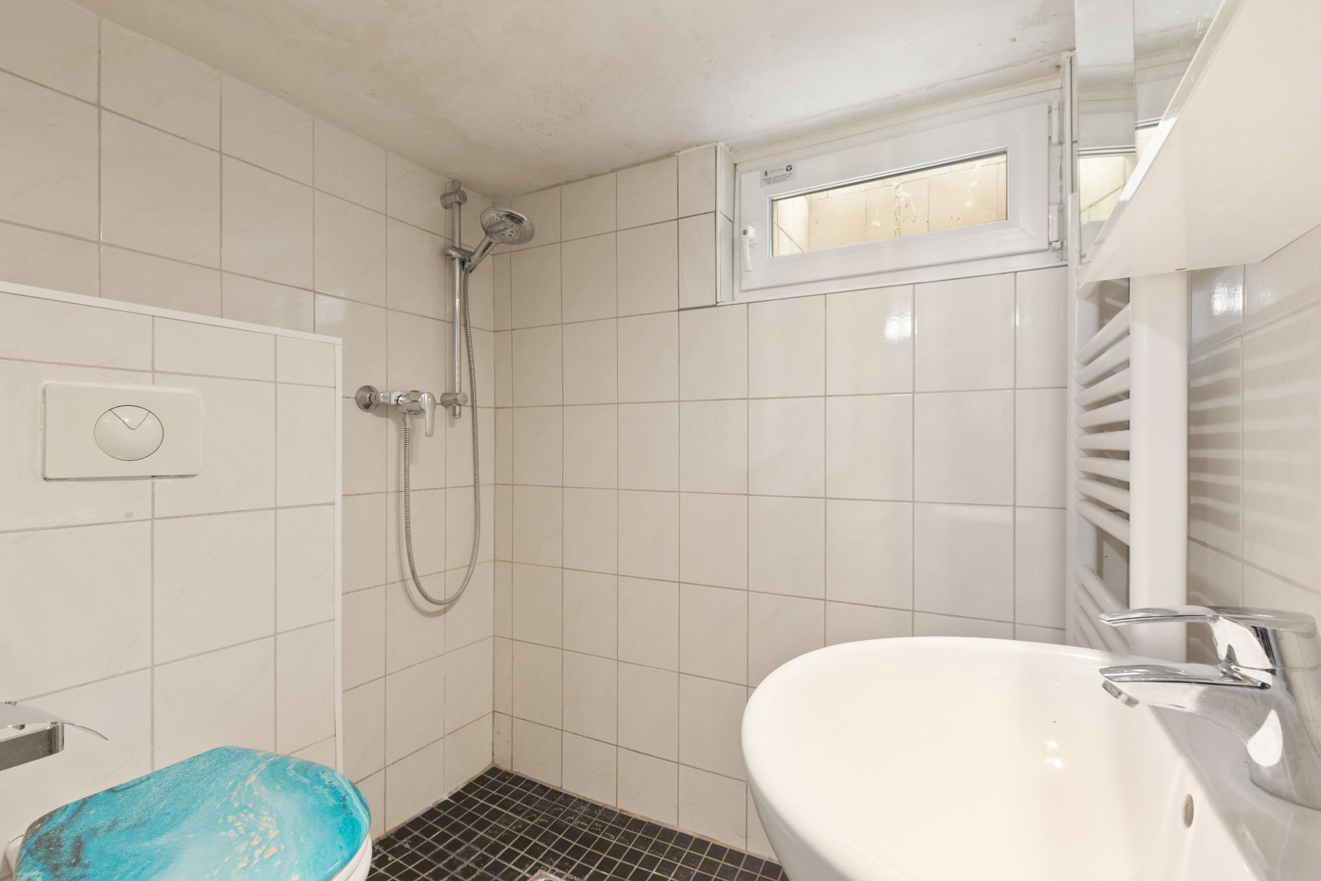 Kleines Bad mit Dusche, Toilette mit blauem Sitz, Waschbecken und kleinem Fenster.