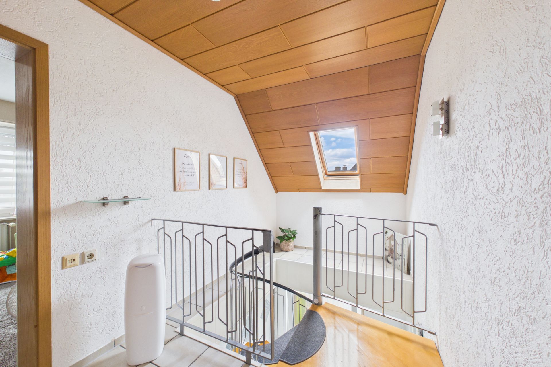 Helle Flur mit Treppengeländer, Dachfenster, Holzdecke und dekorativen Bildern an der Wand.