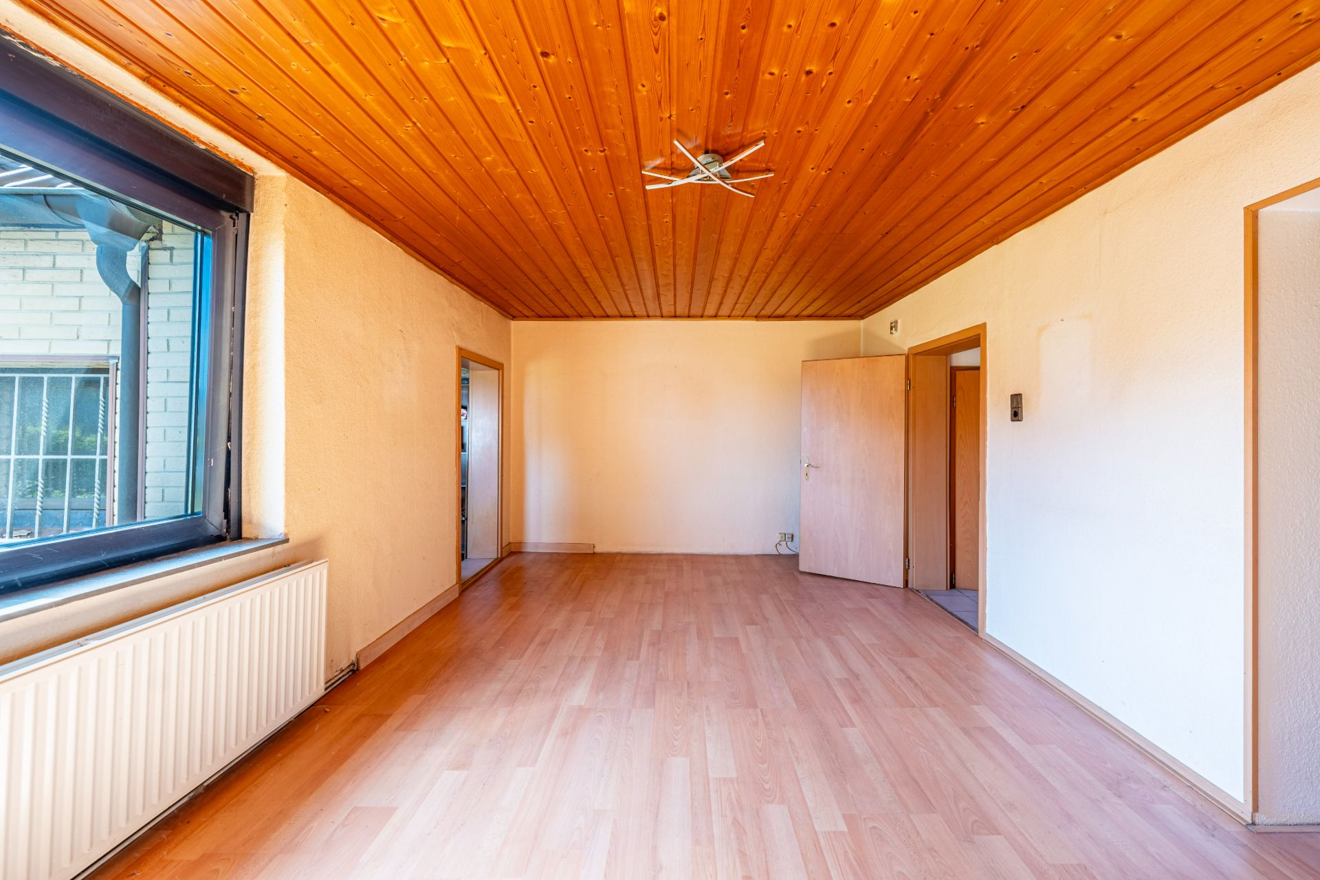Leeres Zimmer mit Holzdecke, großem Fenster, Heizkörper und Holztür.