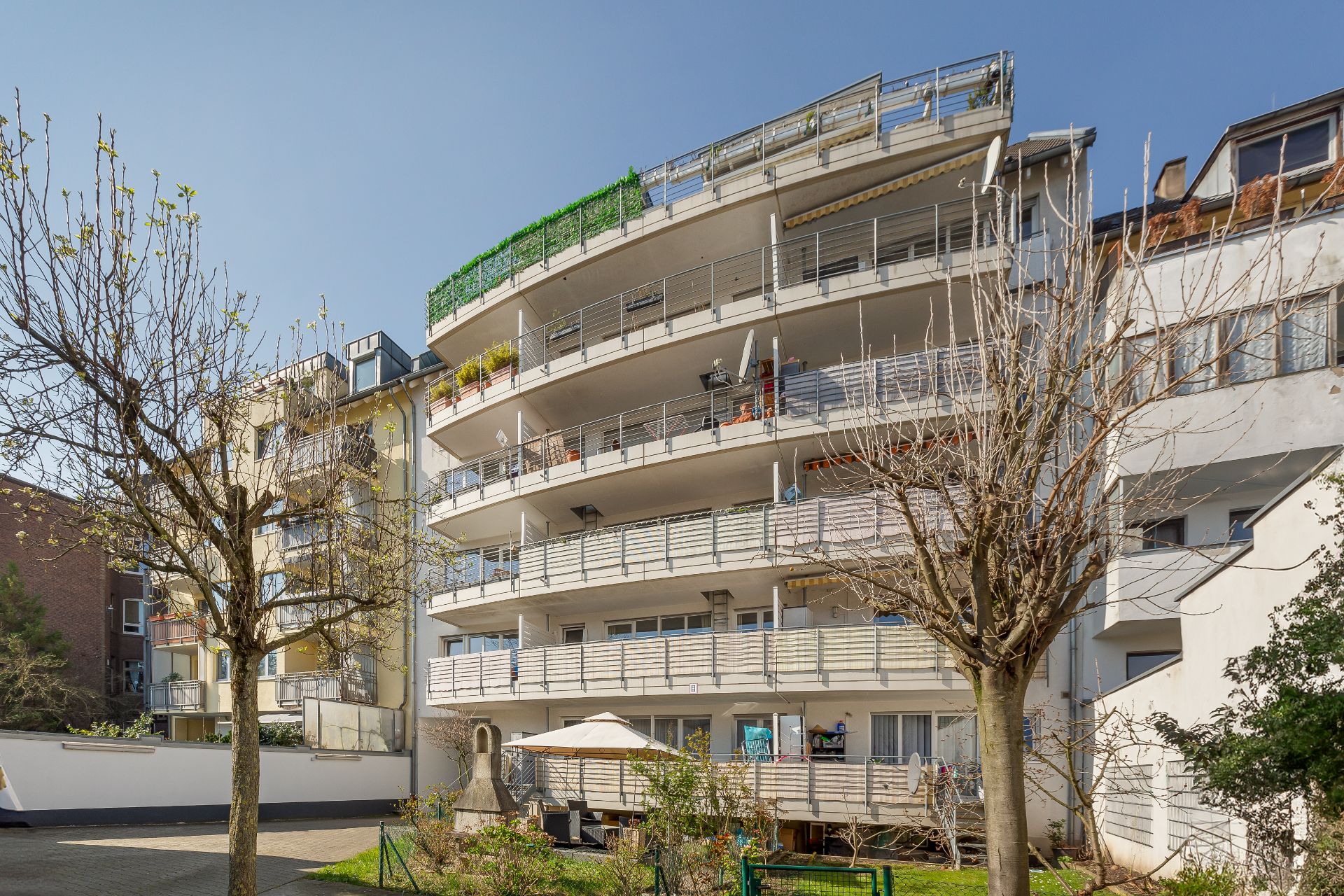 Mehrstöckiges Wohnhaus mit Balkonen und kleinem Gartenbereich vorne.