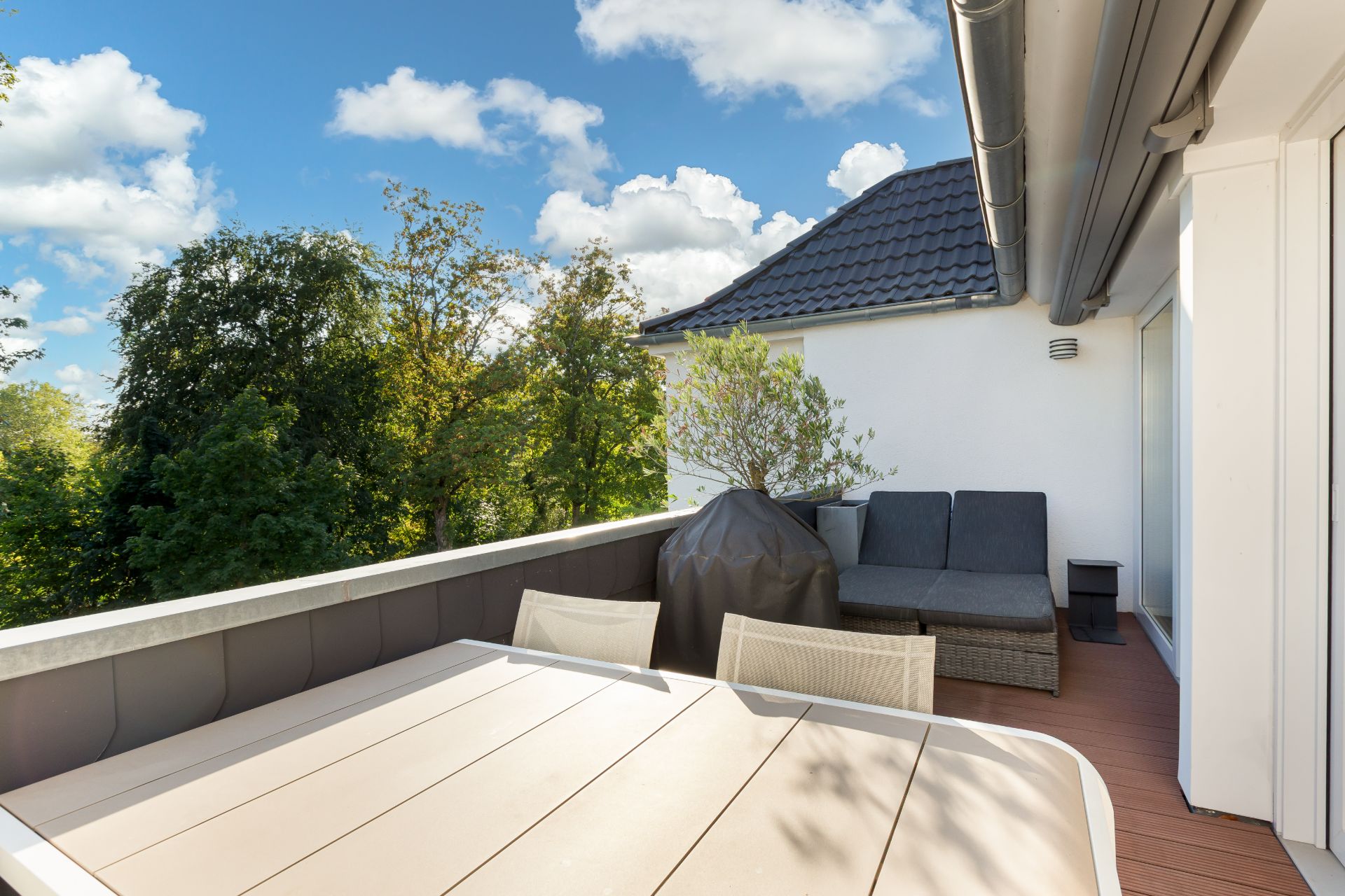 Balkon mit Tisch, Stühlen, Grill, Sofa und Blick auf Bäume an einem sonnigen Tag.