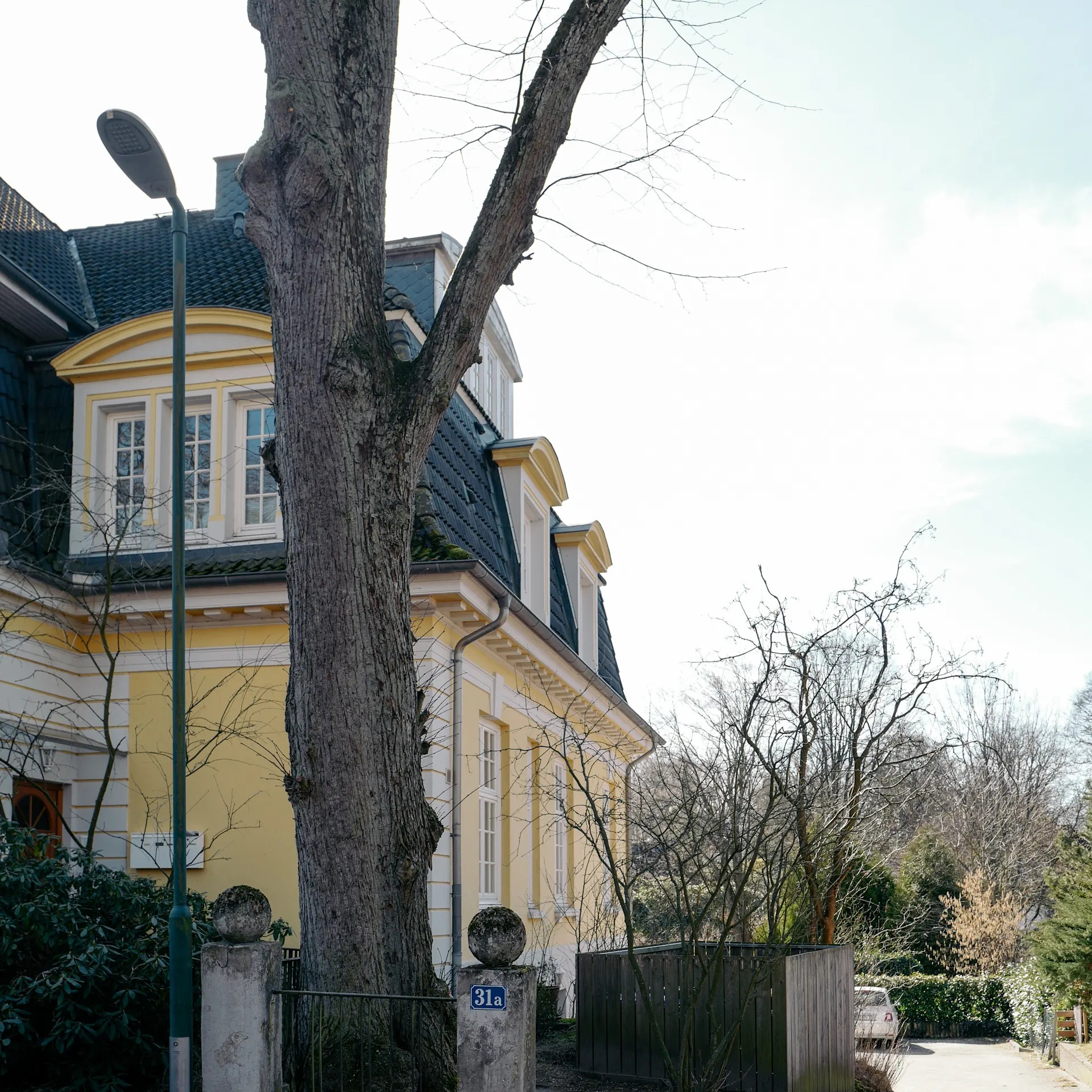 Gelbes Wohnhaus mit schwarzem Dach an einer gepflasterten Straße, Baum und Zaun im Vordergrund.