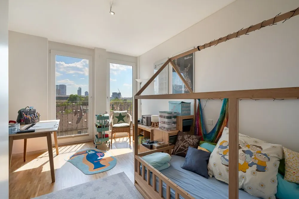 Helles Kinderzimmer mit großem Fenster, Holzbett in Hausform, Schreibtisch und Spielteppich.