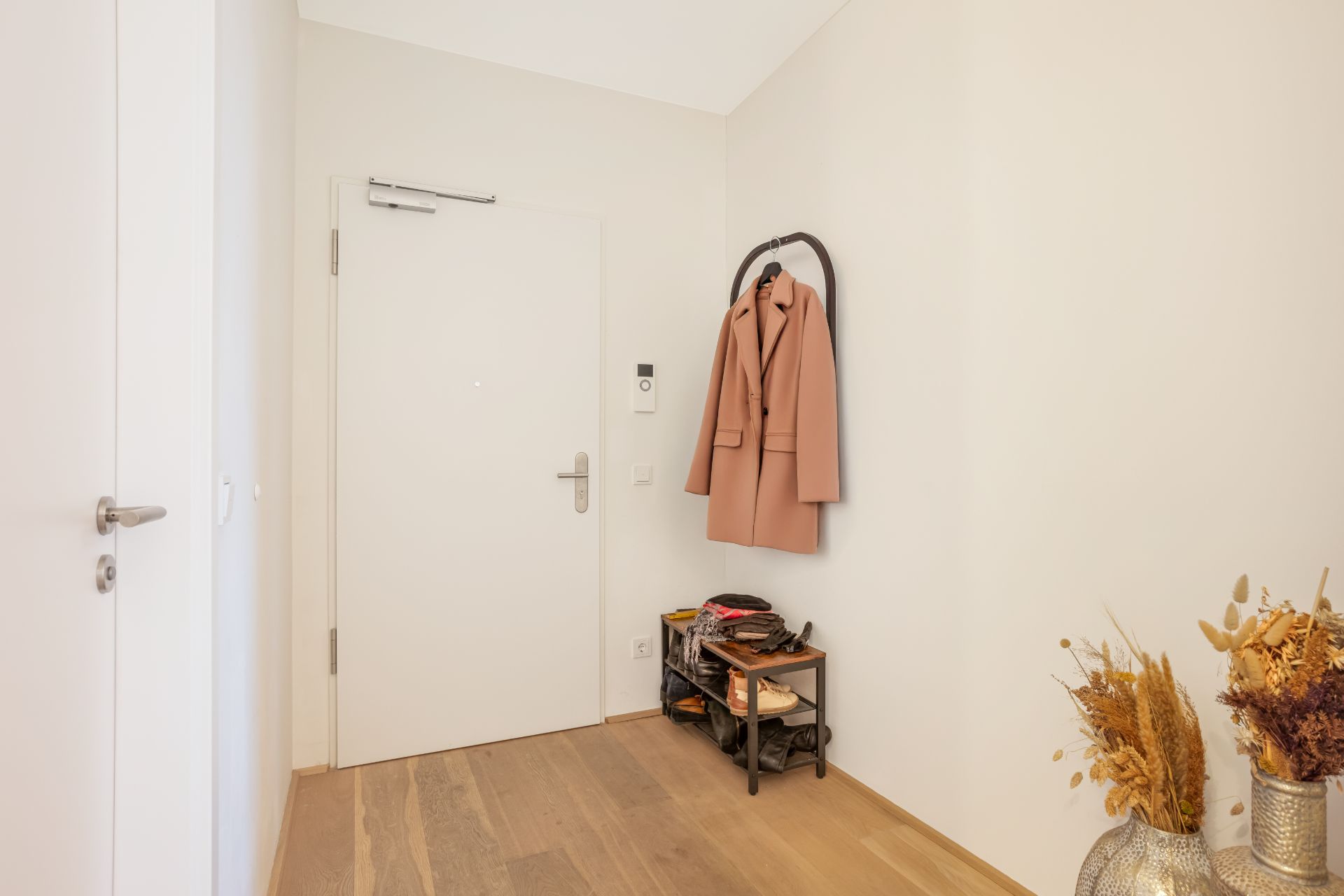 Eingangsbereich mit weißer Tür, Garderobe mit Mantel, Regal und Trockenblumen in einer Vase.