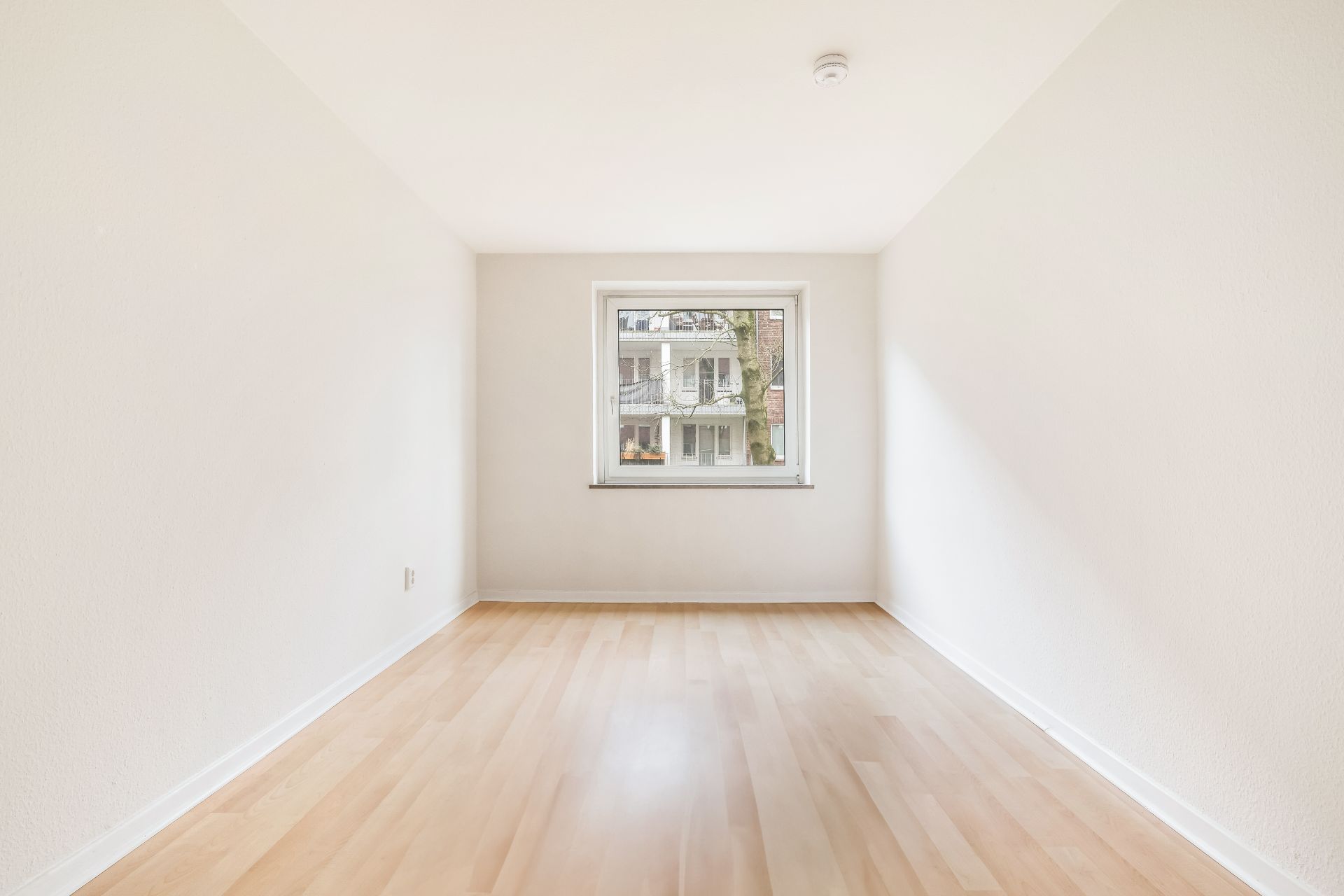 Helles leeres Zimmer mit Holzfußboden und Fenster, das auf ein Nachbargebäude blickt.