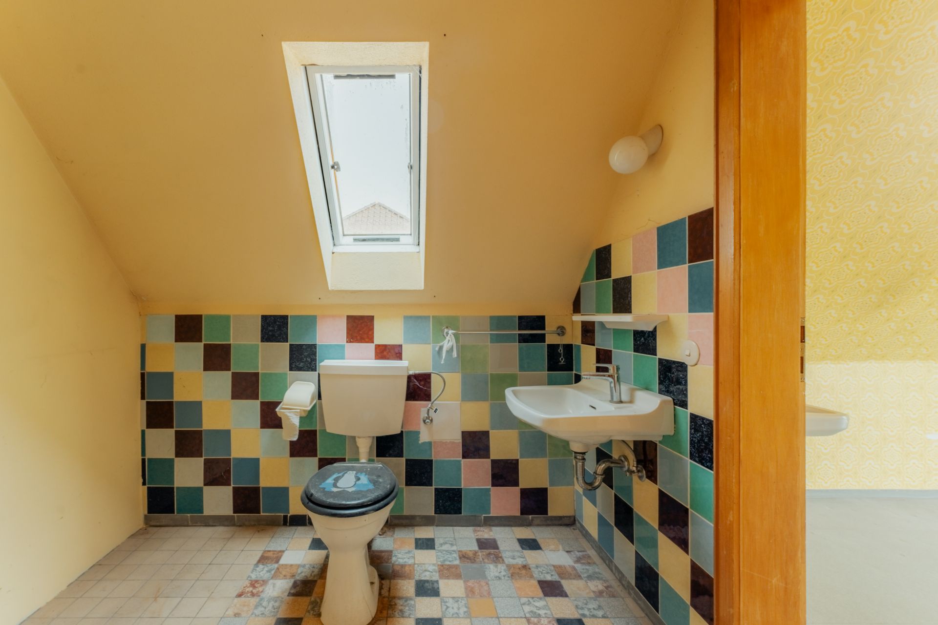Kleines Badezimmer mit Dachfenster, Toilette, Waschbecken und bunten Wandfliesen.