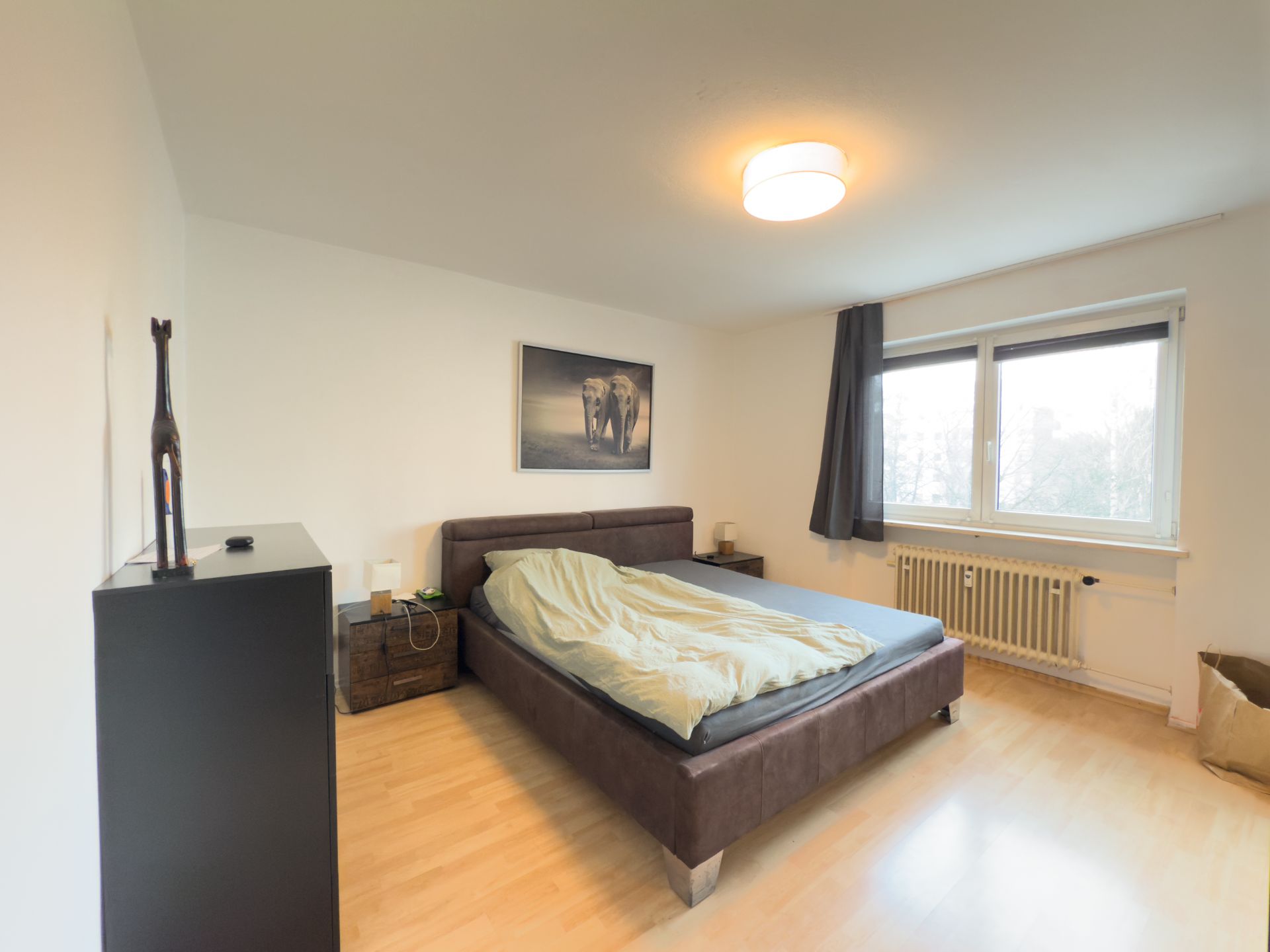 Schlafzimmer mit Doppelbett, Nachttischen, großem Fenster und Kommode auf Parkettboden.