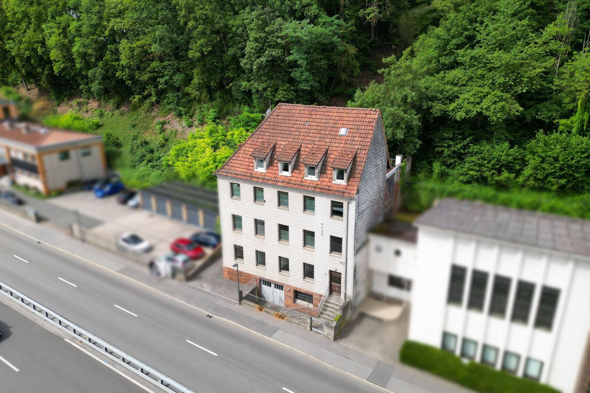Dreistöckiges Wohngebäude mit rotem Dach an einer Straße, umgeben von Bäumen und Nachbarhäusern.