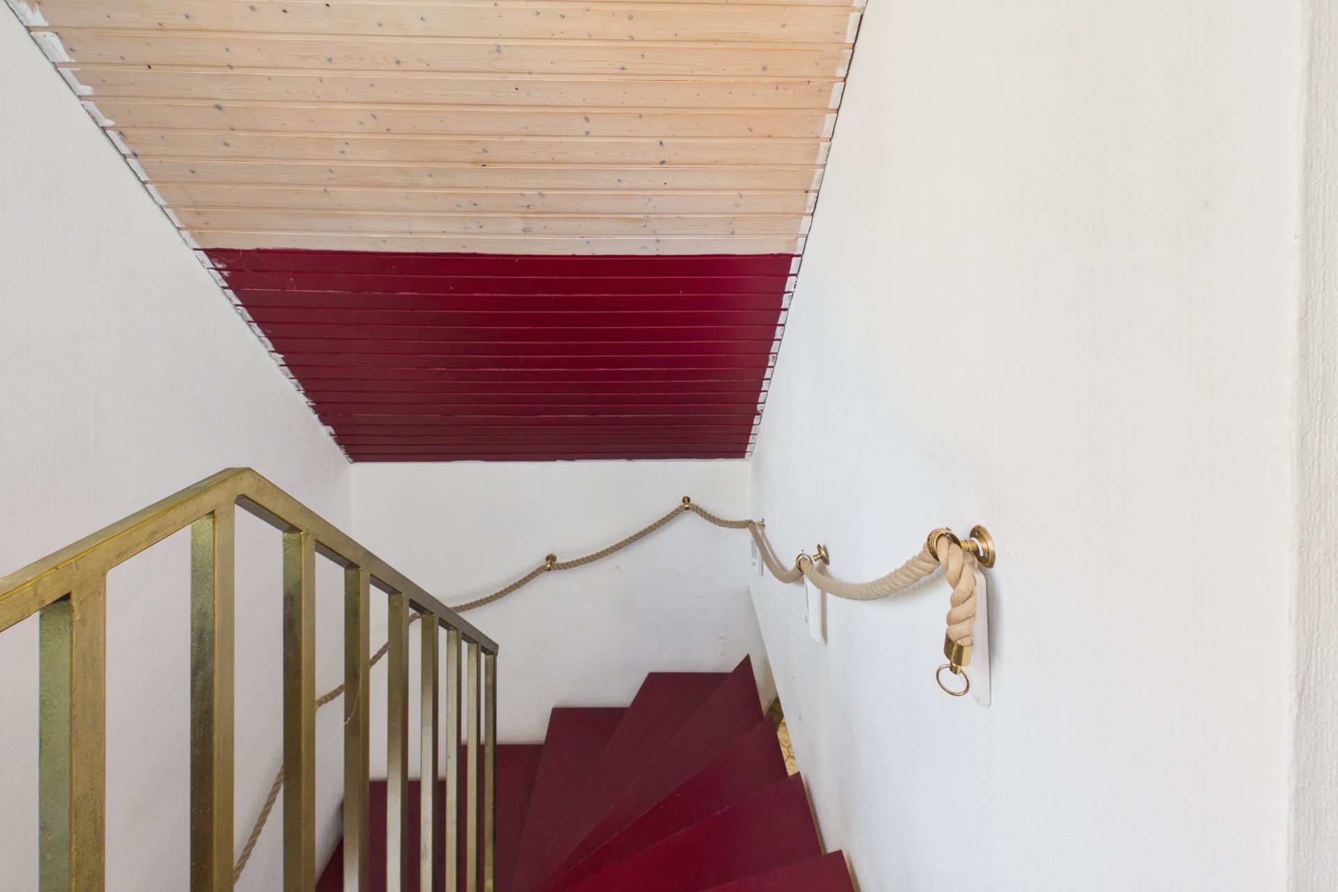 Rote Treppe mit goldenem Geländer und Seil-Handlauf in einem weißen Treppenhaus.