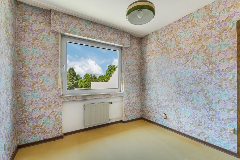 Leeres Zimmer mit bunter Blumen-Tapete, Fenster und Heizkörper unter dem Fenster.