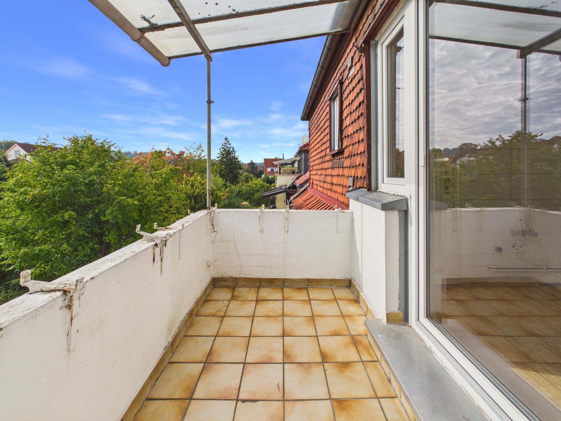 Überdachter Balkon mit Fliesenboden und Blick auf grüne Bäume, angrenzend an ein Haus mit roten Dachschindeln.