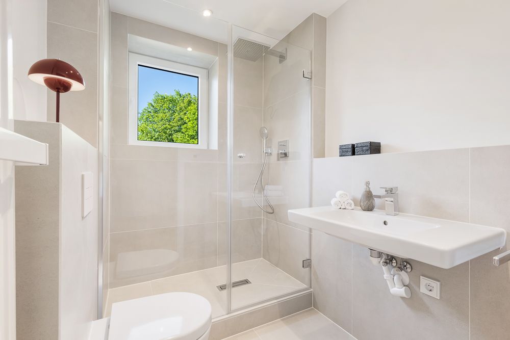 Modernes Badezimmer mit Dusche, Waschbecken, WC und Fenster mit Blick auf grünen Baum.