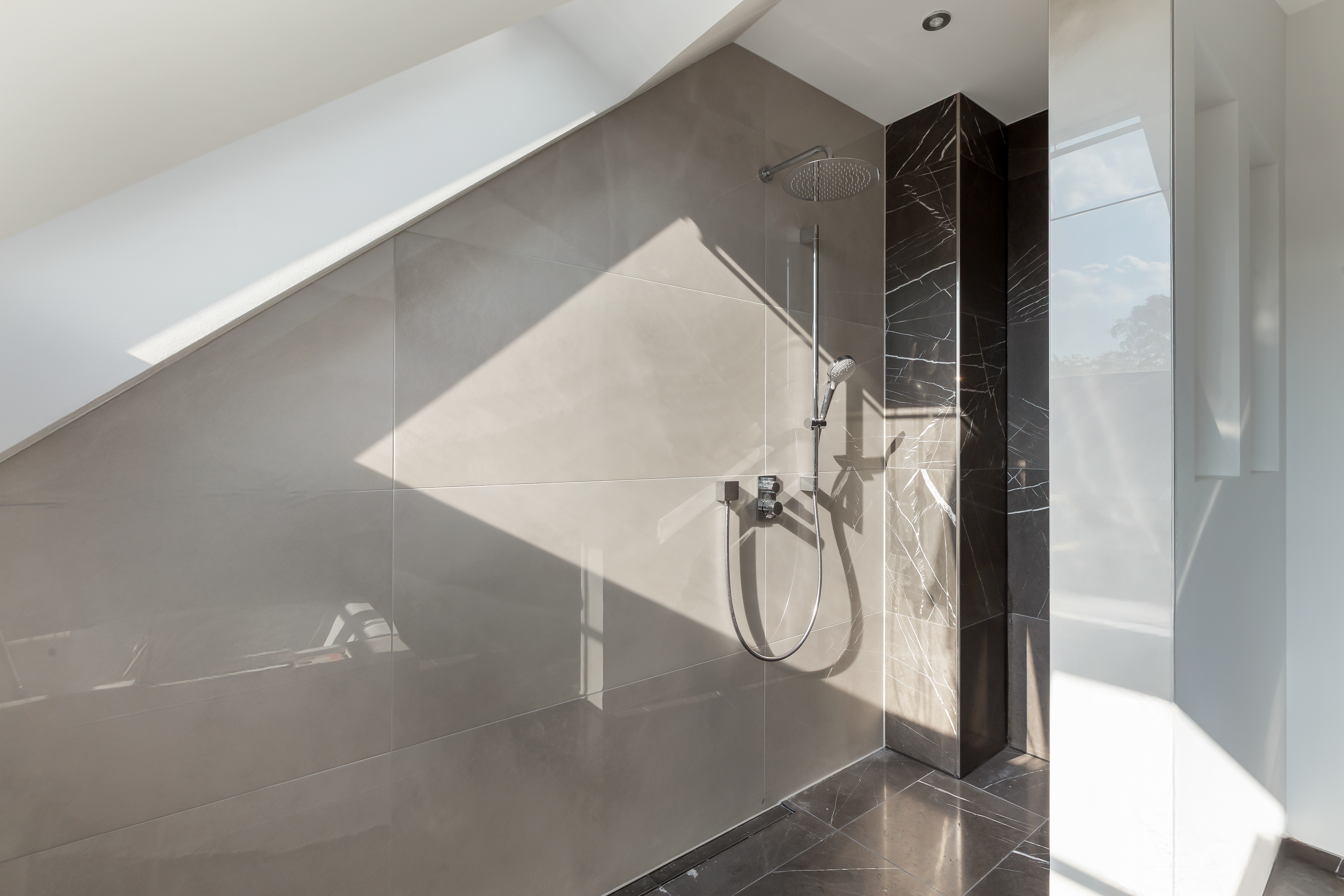 Moderne, offene Dusche mit grauen Fliesen und schräger Decke im Tageslicht.