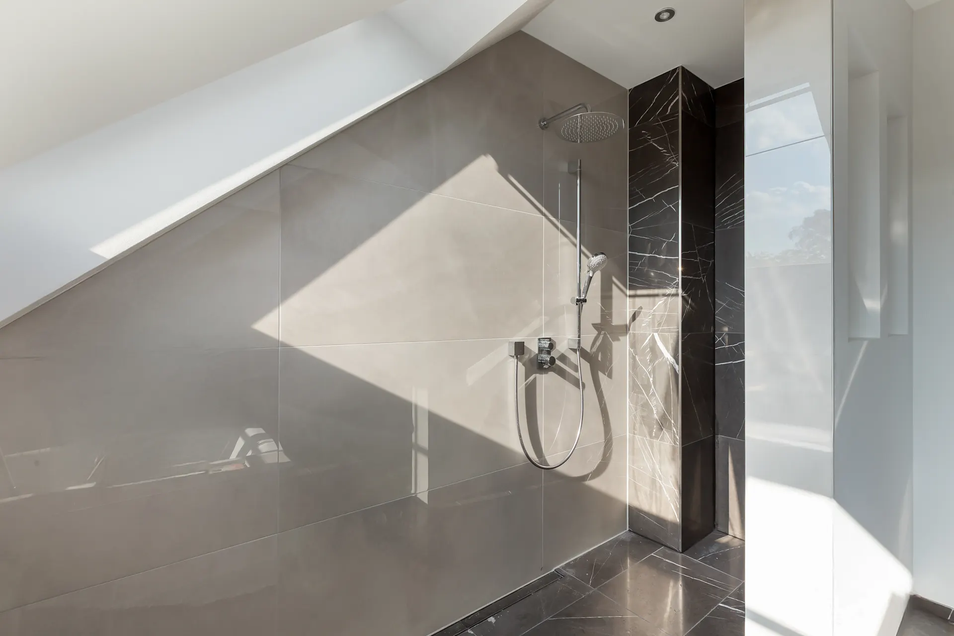 Moderne, offene Dusche mit grauen Fliesen und schräger Decke im Tageslicht.