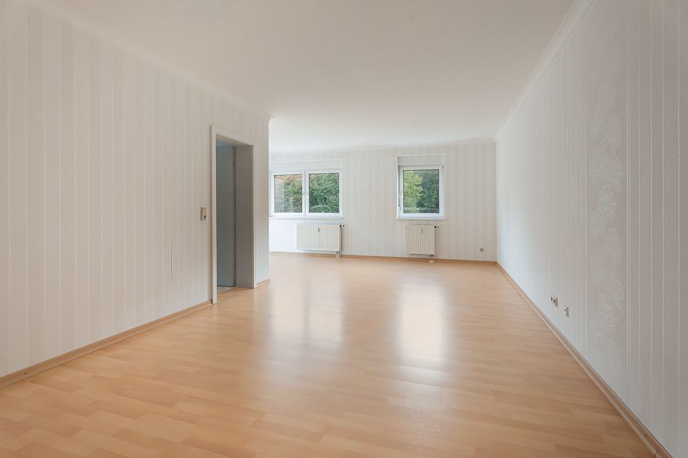 Leeres Wohnzimmer mit hellem Laminatboden, weißen Wänden und zwei Fenstern.
