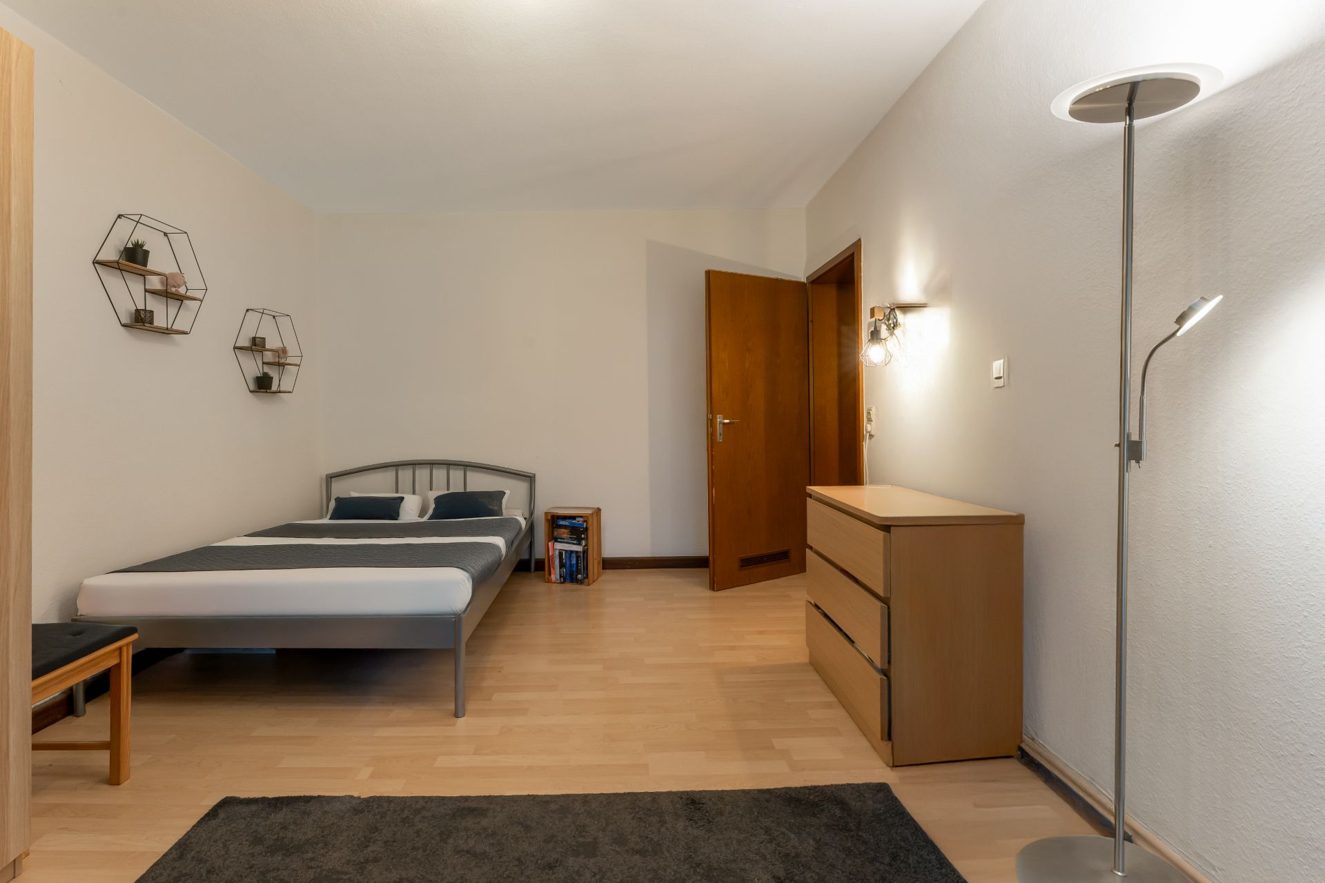 Schlafzimmer mit Doppelbett, Kommode, Stehlampe und offenem Holztür, helle Wände und Holzboden.