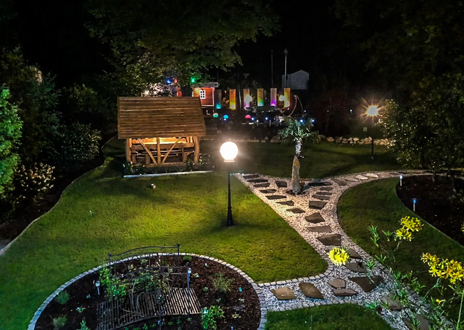 Nachts beleuchteter Garten mit Rasen, Wegen aus Steinen, Pavillon und Blumenbeeten.