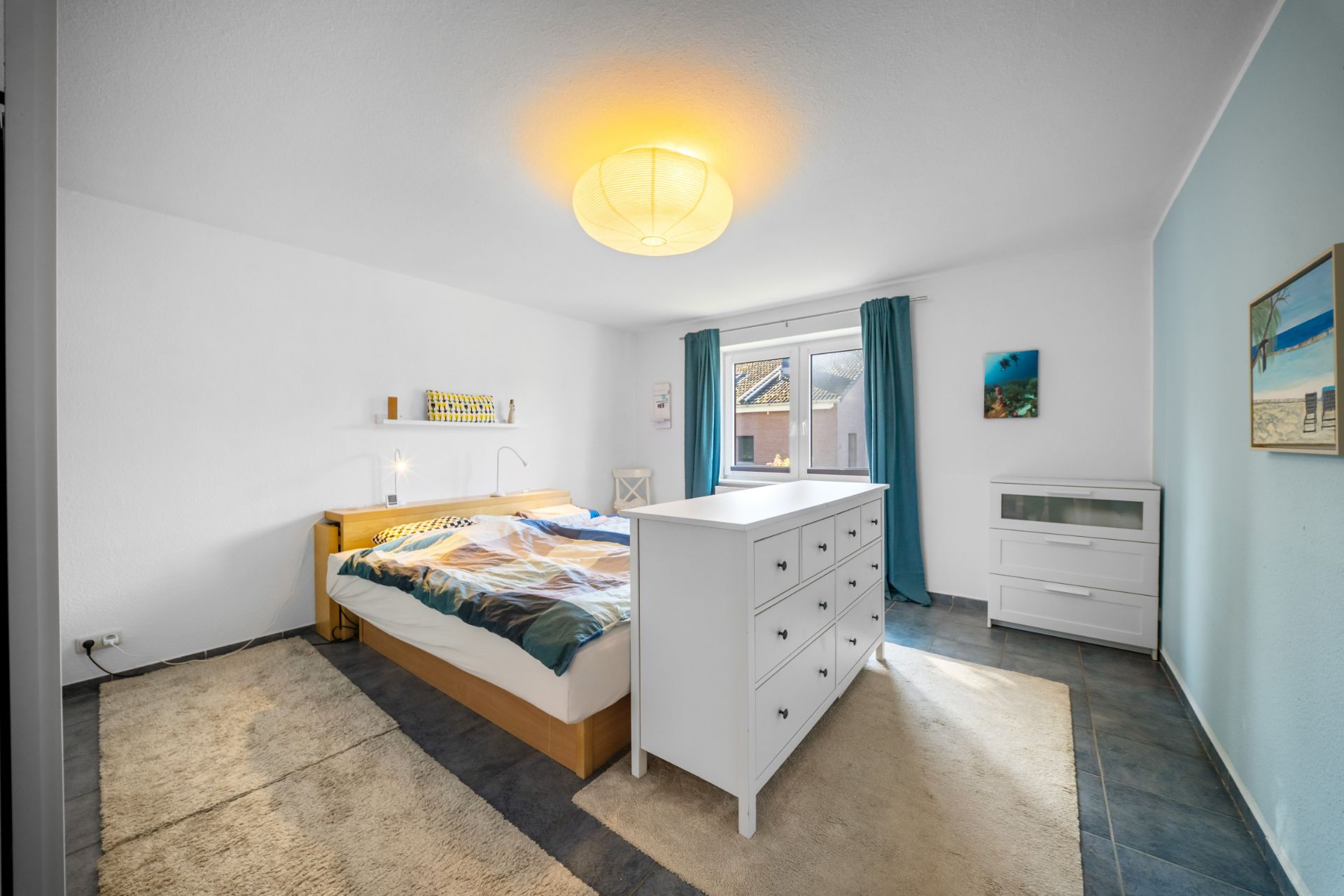 Helles Schlafzimmer mit Doppelbett, weißer Kommode, Teppich und Fenster mit blauen Vorhängen.