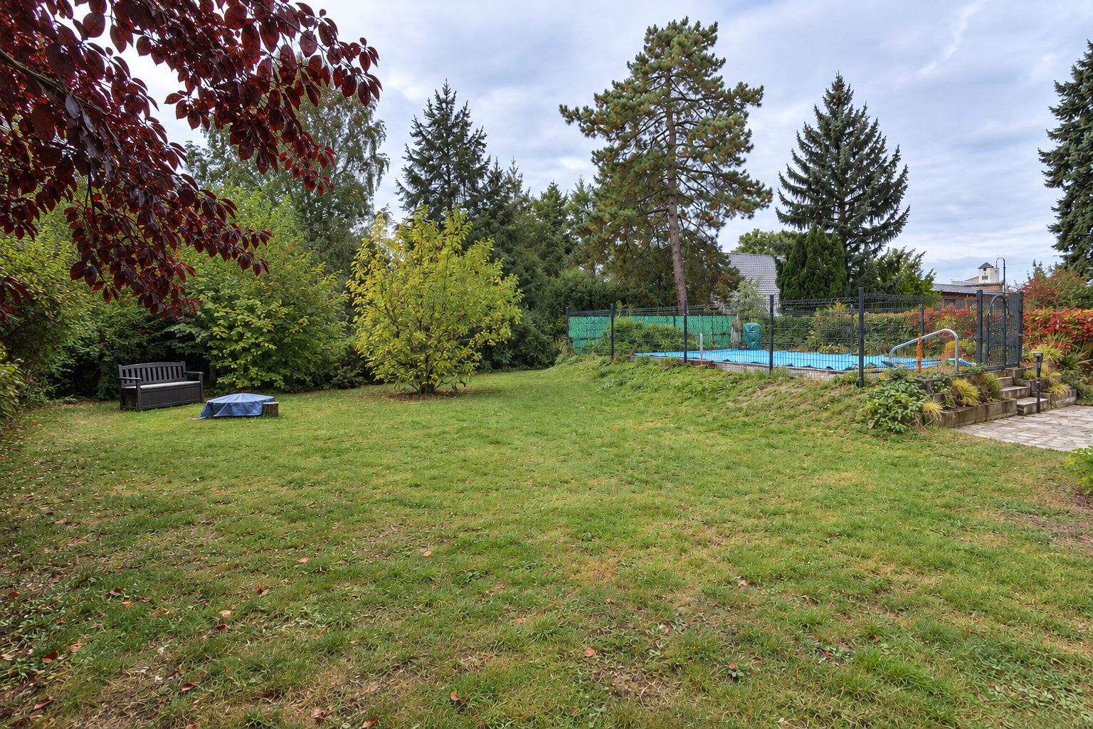 Großer Garten mit Rasen, Bäumen, einer Bank und eingezäuntem Poolbereich rechts.