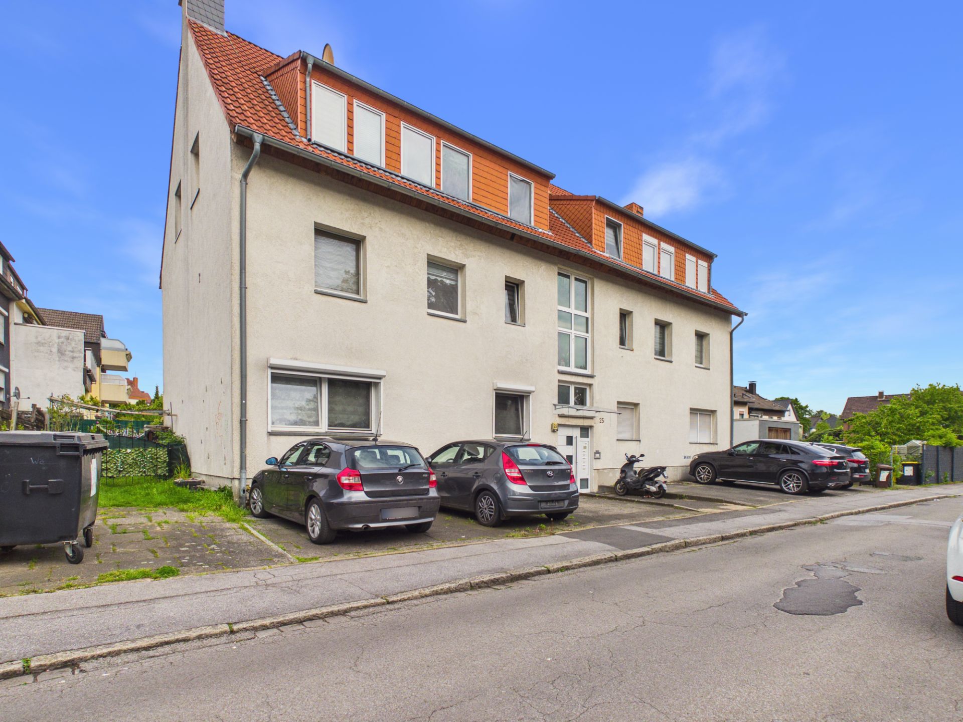 Mehrfamilienhaus mit mehreren geparkten Autos vor dem Eingang an einer Wohnstraße.